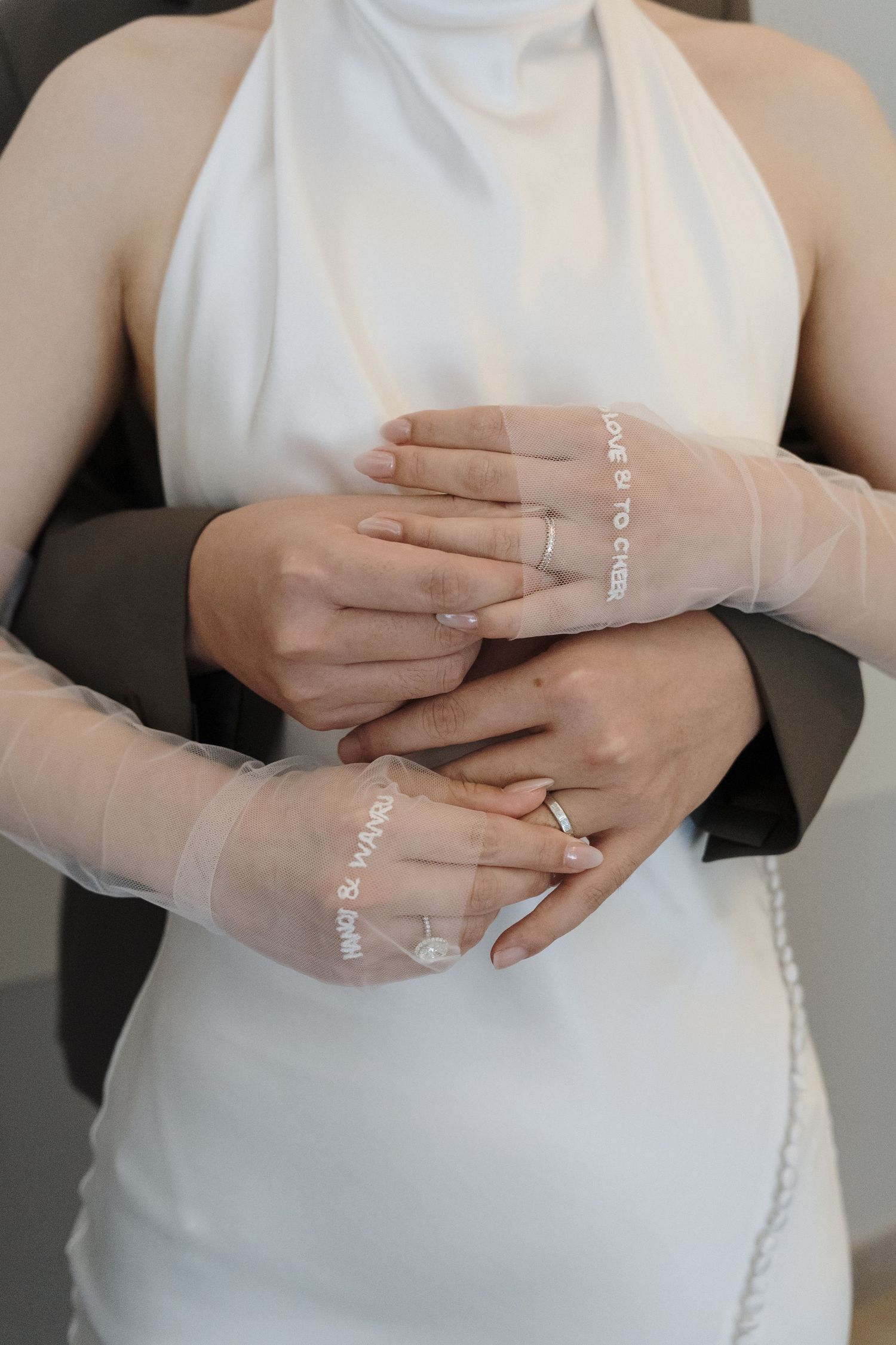 Bride and groom holding hands showing wedding rings, with sheer embroidered gloves and halter gown for editorial wedding photography.