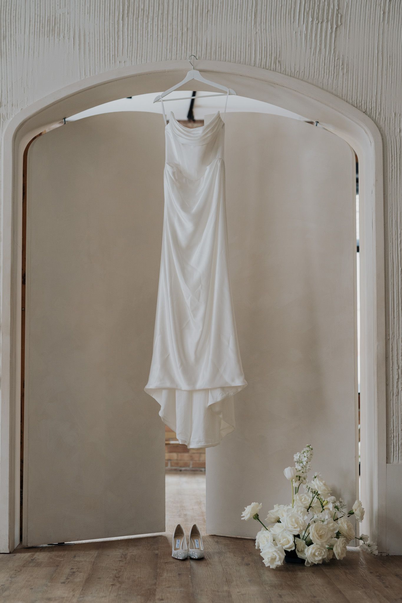 White satin wedding dress hanging in an arched doorway with bridal heels and bouquet at Archive Studio in Toronto editorial wedding