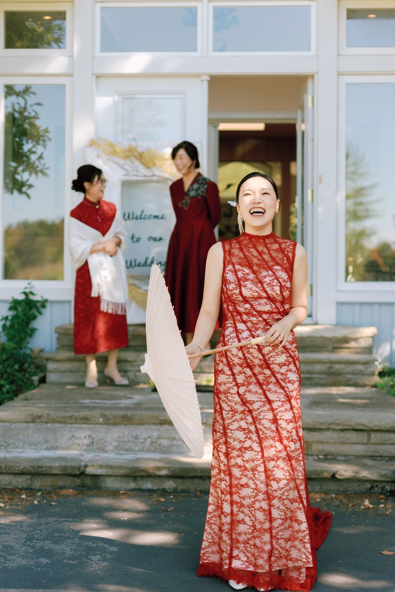 Laughing woman in red lace qipao holding a white fan during a Chinese tea ceremony wedding at an estate venue with guests behind