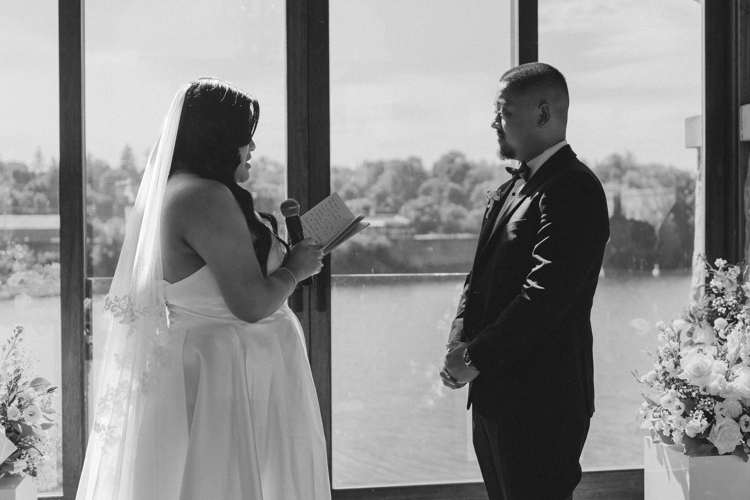 A bride reads her vows to her groom in a black and white waterfront wedding ceremony photo.