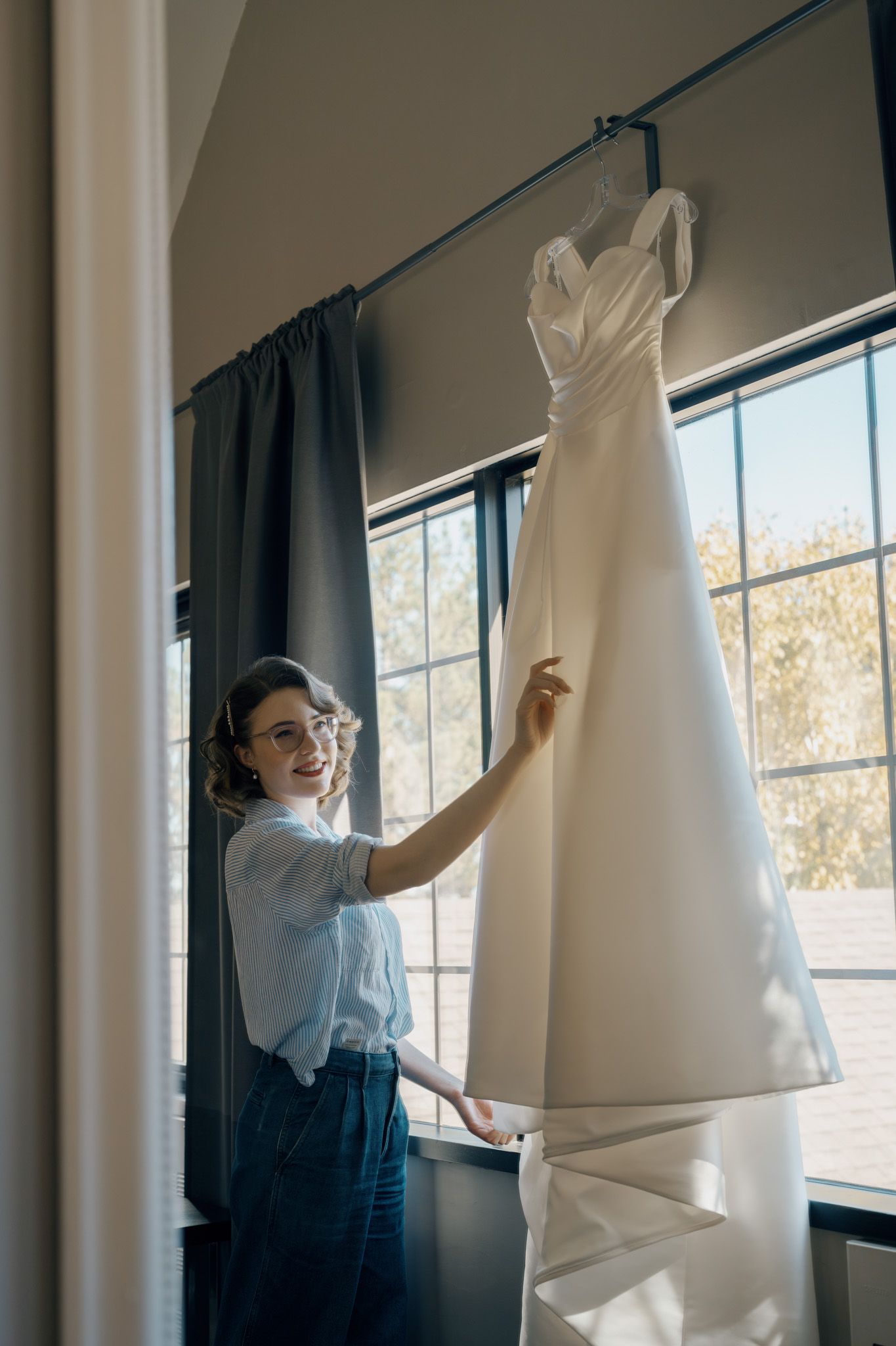 Laughing bride holding her white wedding gown by a bright window at The Manor in King City during her wedding morning.