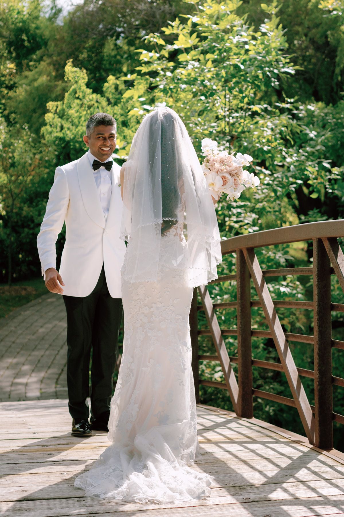 Groom in white tuxedo walking toward bride in lace gown on a garden bridge at Kurtz Orchards in Niagara-on-the-Lake.