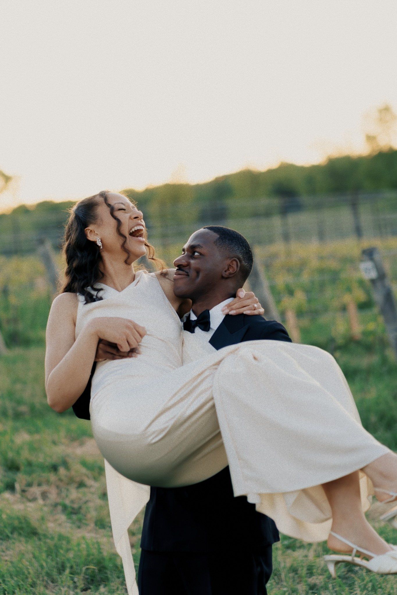Joyful newlyweds at vineyard sunset, groom in tuxedo carrying laughing bride in white wedding dress.