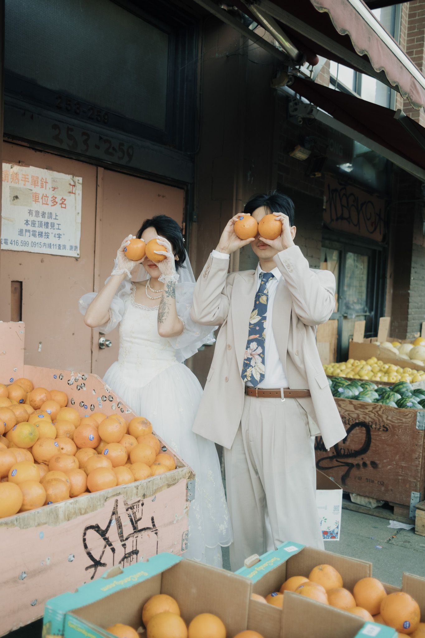 Couple in wedding attire holding oranges at a fruit stand on Chinatown, Toronto during a Korean-style pre-wedding shoot