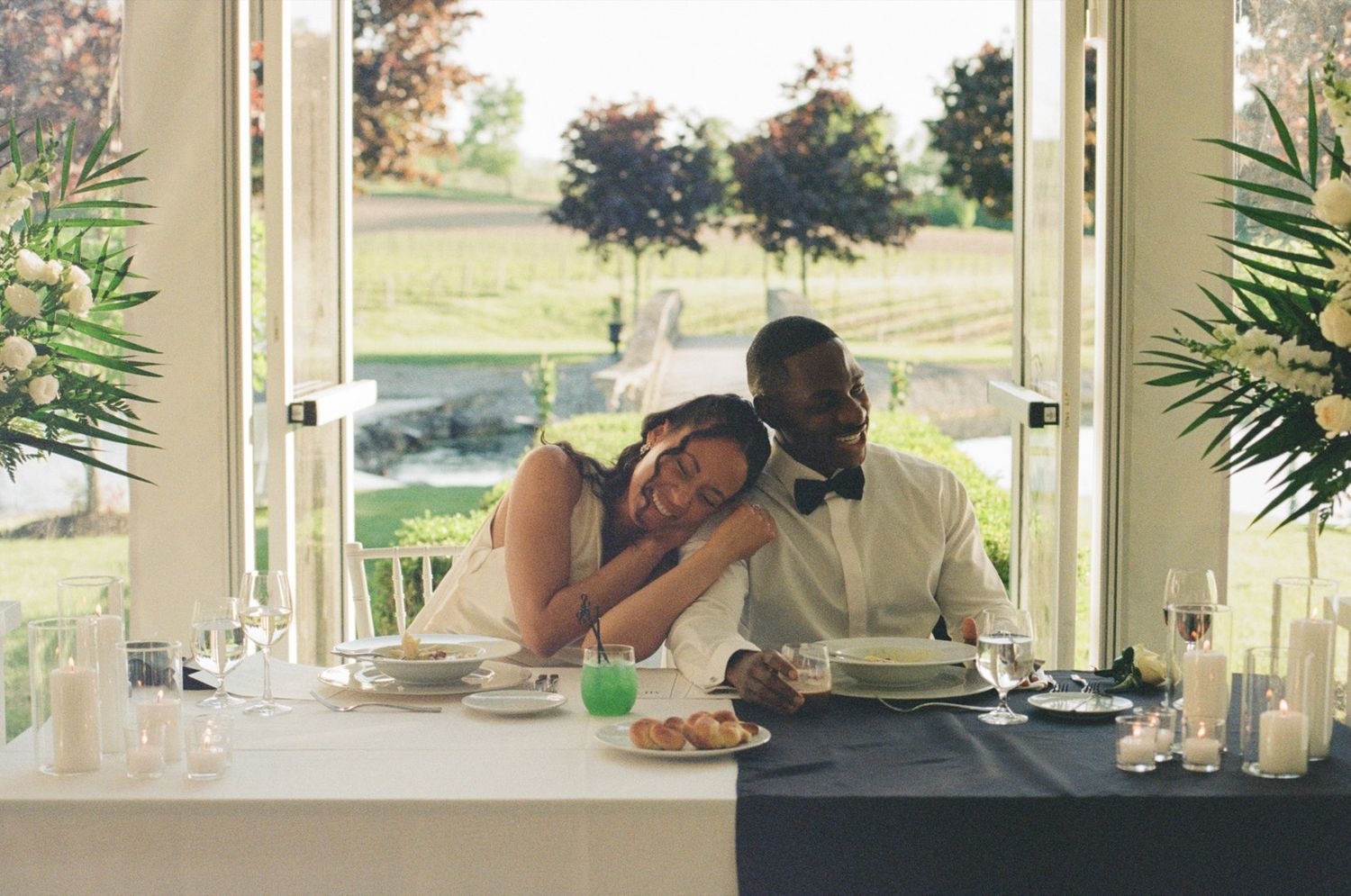 Bride resting head on groom's shoulder at wedding reception table with candles and green garden view.