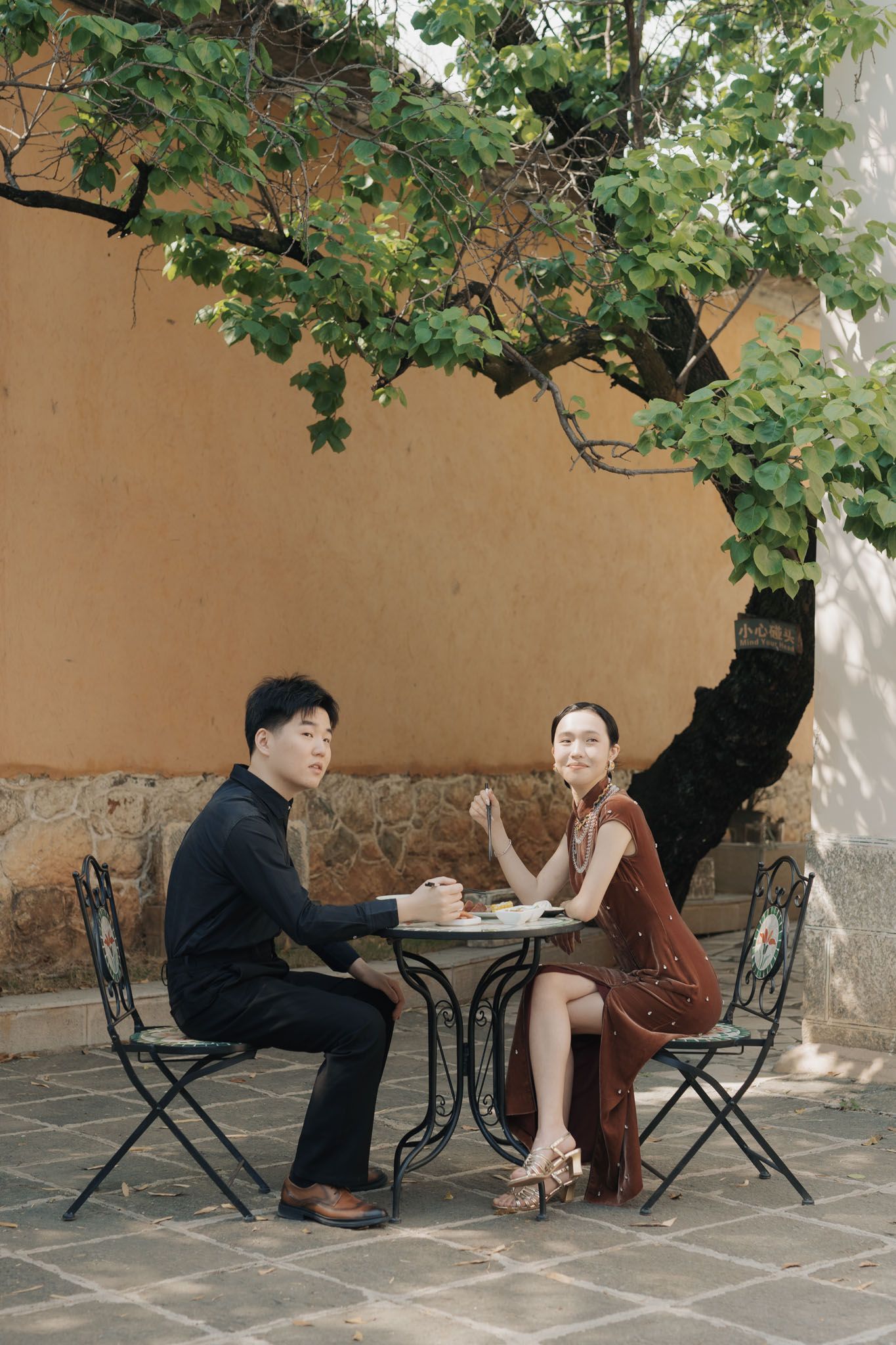 Couple seated at an outdoor café beneath a green tree and yellow wall during their destination wedding portraits.