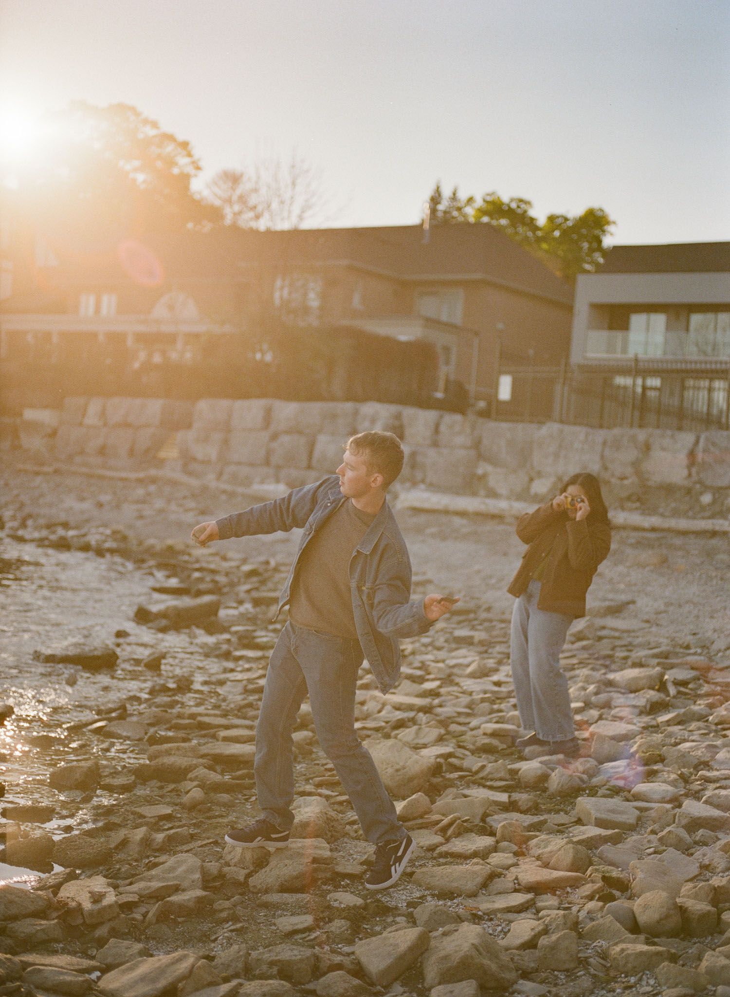 Couple playing on a rocky shoreline at golden hour with sunlit buildings in the background. Mississaugua beach sunset portraits