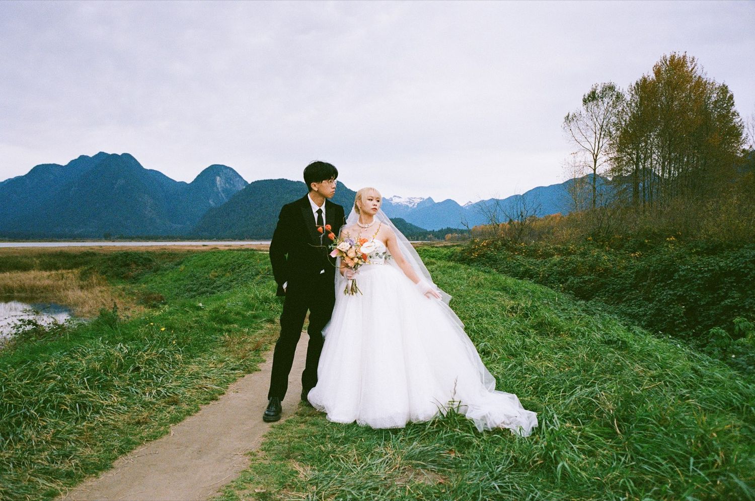 A bride and groom walk a grassy path with mountains and a lake backdrop on their wedding day.