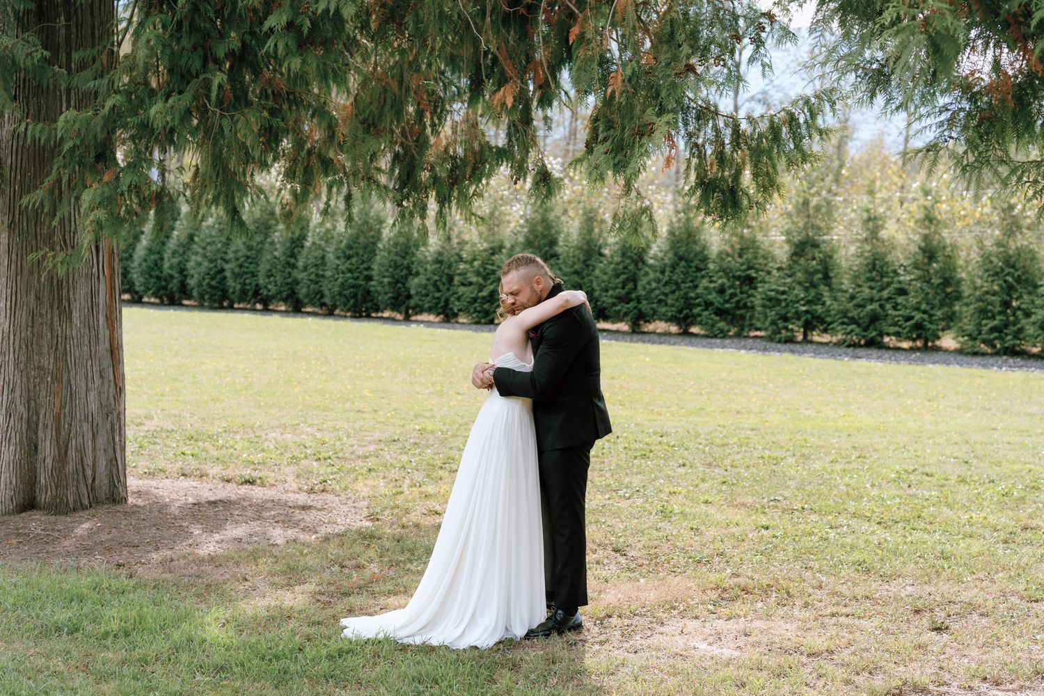 Bride and groom share a romantic embrace under a large evergreen tree at an outdoor wedding.