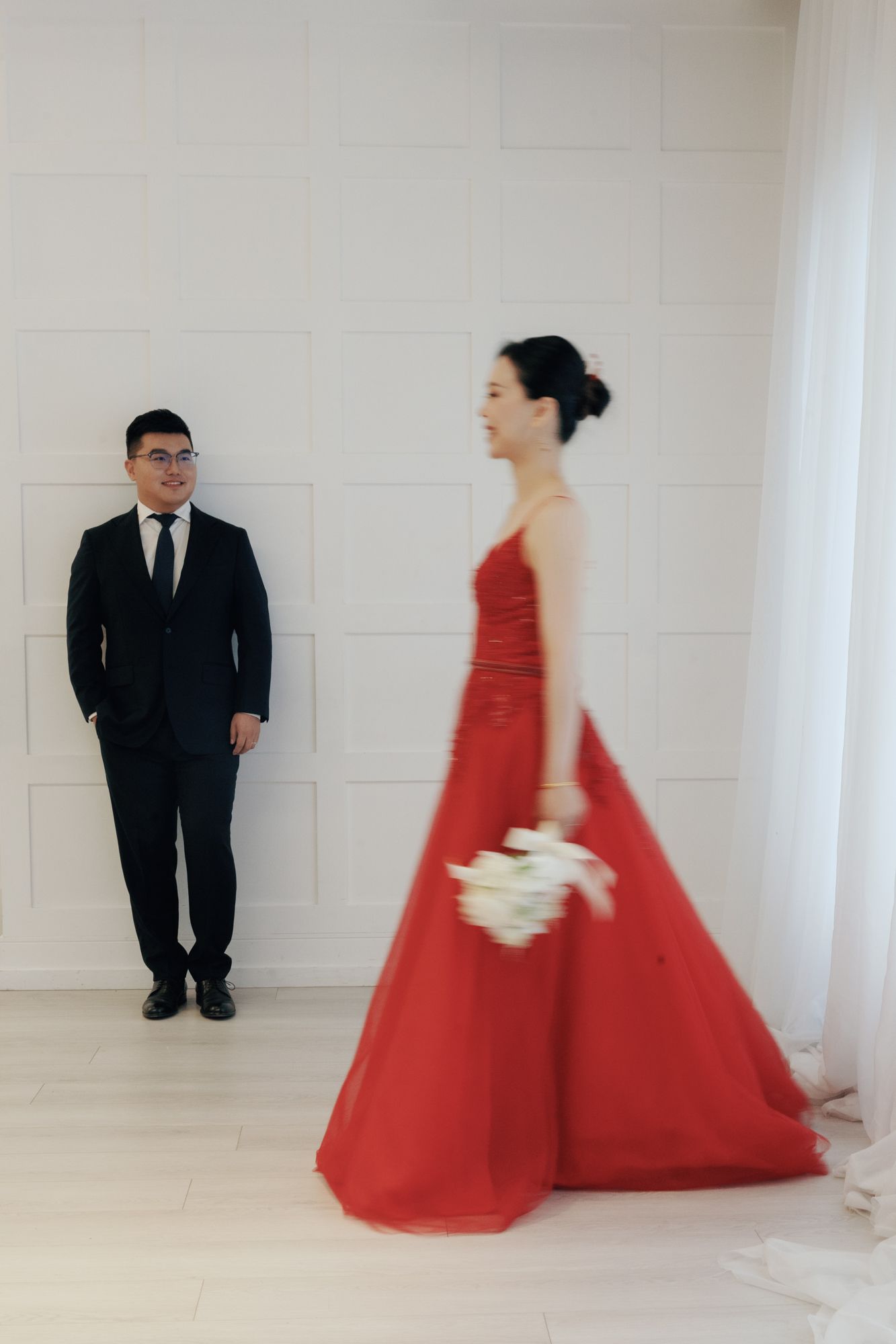 Bride in red gown holding white bouquet as groom watches admiringly in Bellamy Loft North York Toronto.