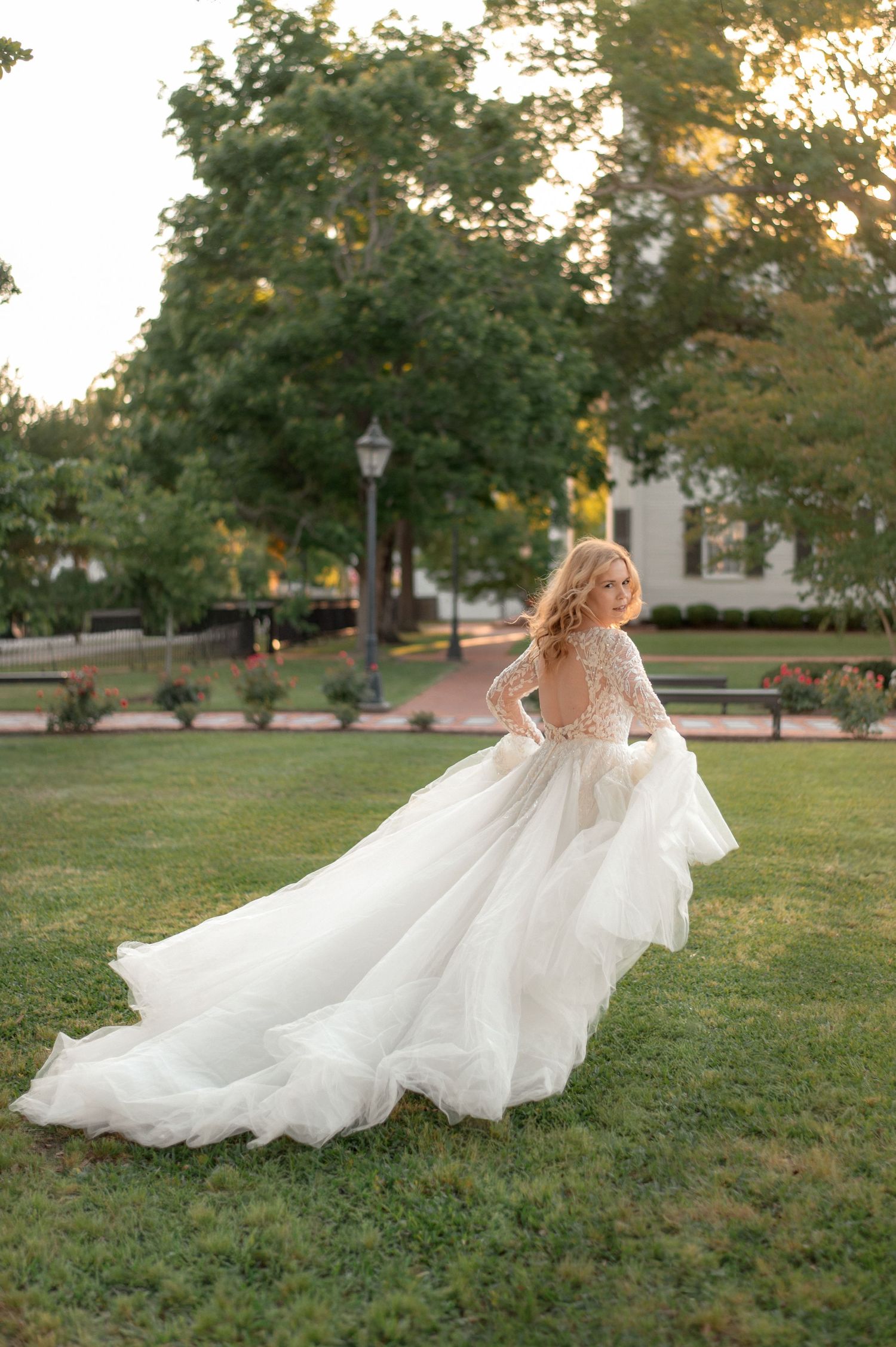 Bride in a lace long-sleeve ballgown with flowing train poses gracefully on green lawn at golden hour.