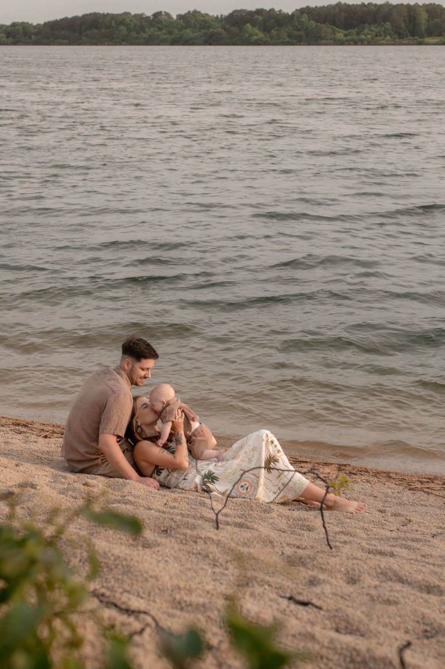 A couple sits together on a sandy beach shore, sharing a tender moment with their baby near calm water.