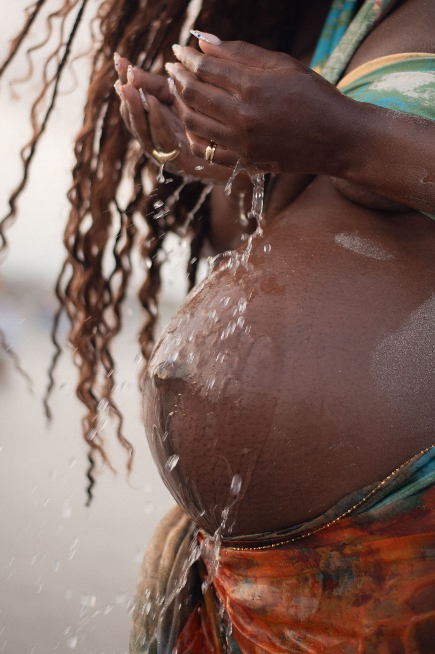 Pregnant woman pouring water over her bare belly outdoors, with curly hair and colorful wrap skirt.