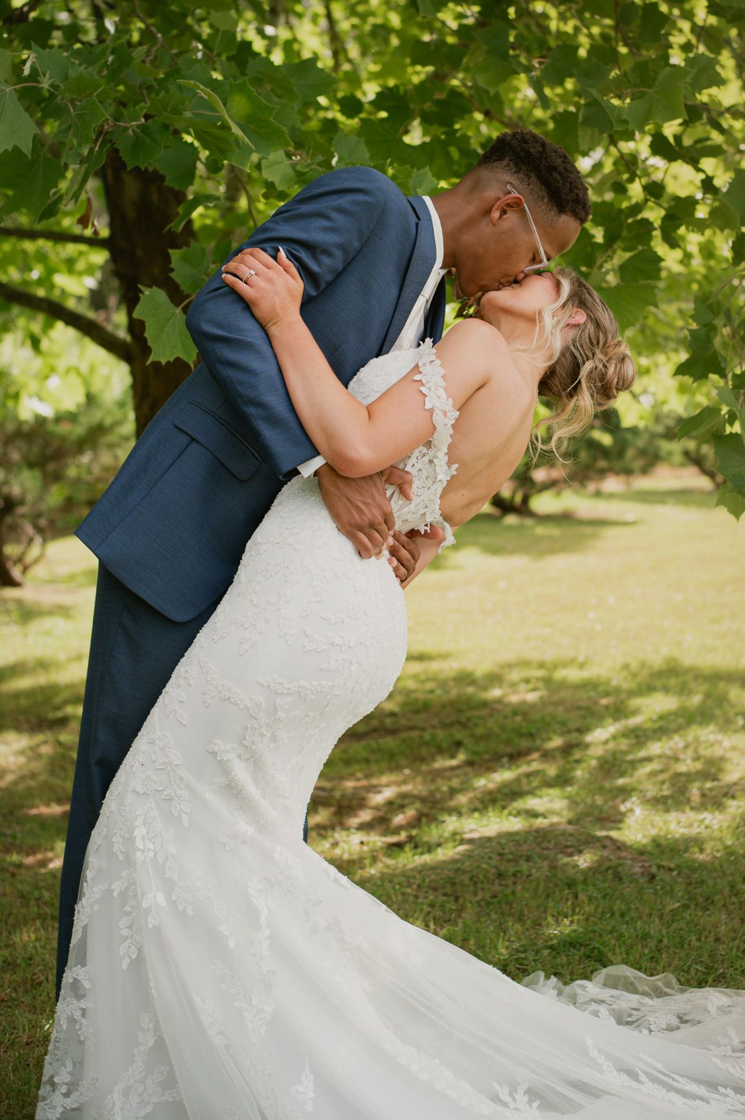 Groom in navy suit dips bride in lace mermaid gown for romantic kiss under green trees outdoors.