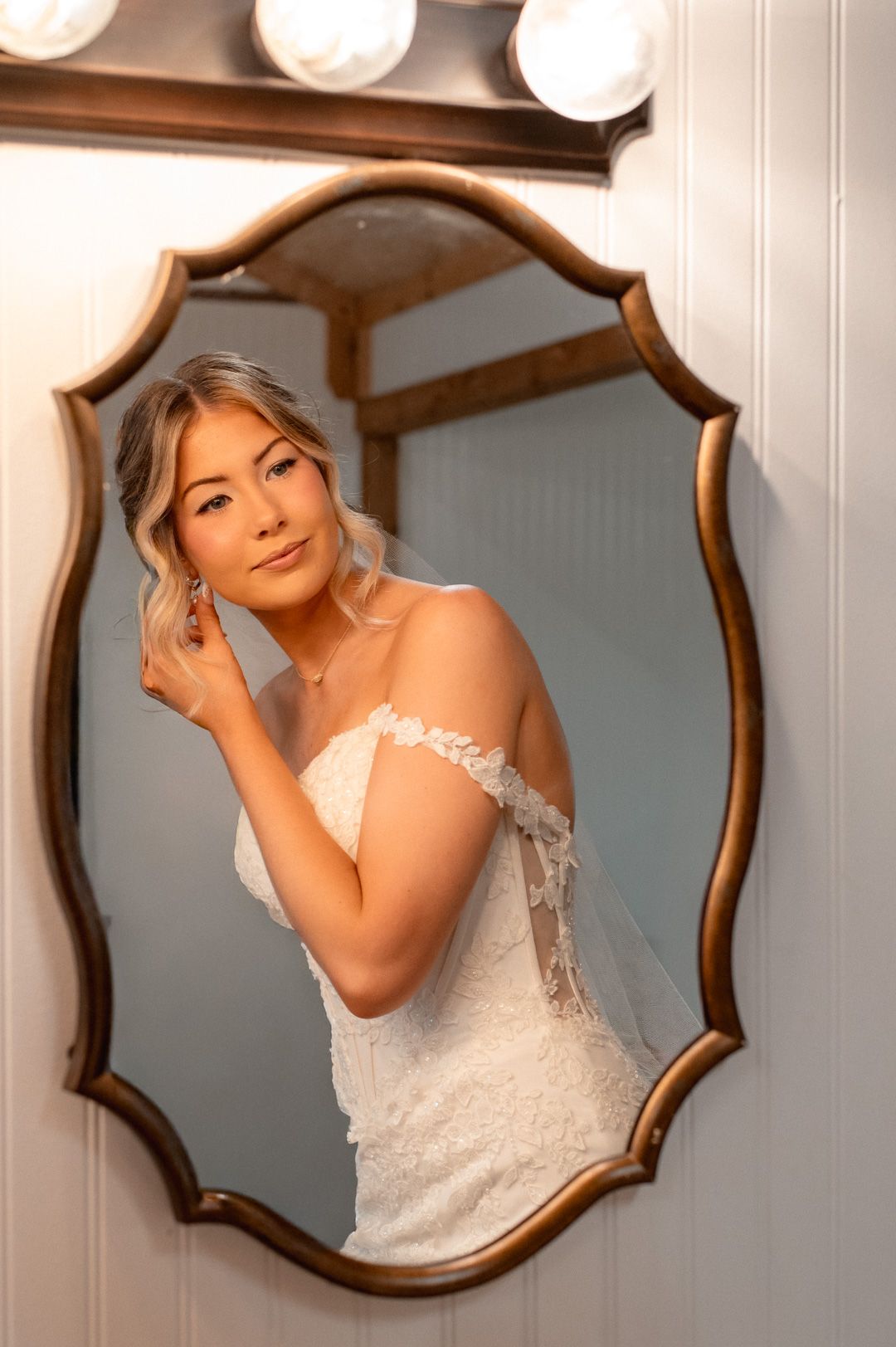 Bride in lace wedding dress and veil adjusting earring, reflected in ornate gold mirror with vanity lights.