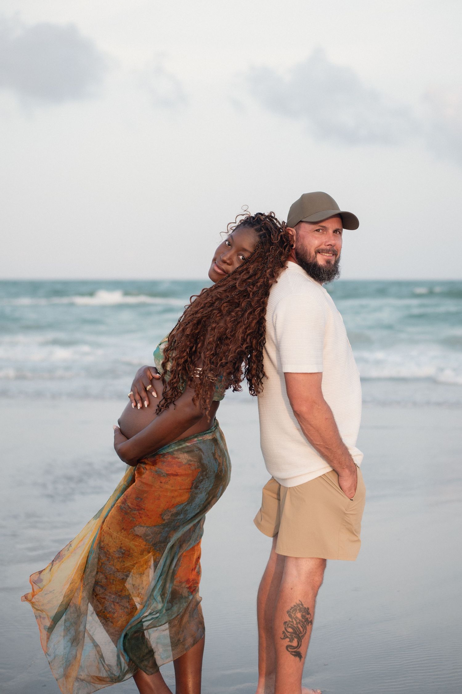 Interracial couple posing back-to-back on a beach, woman in flowing dress with long curly hair, man in cap and shorts.