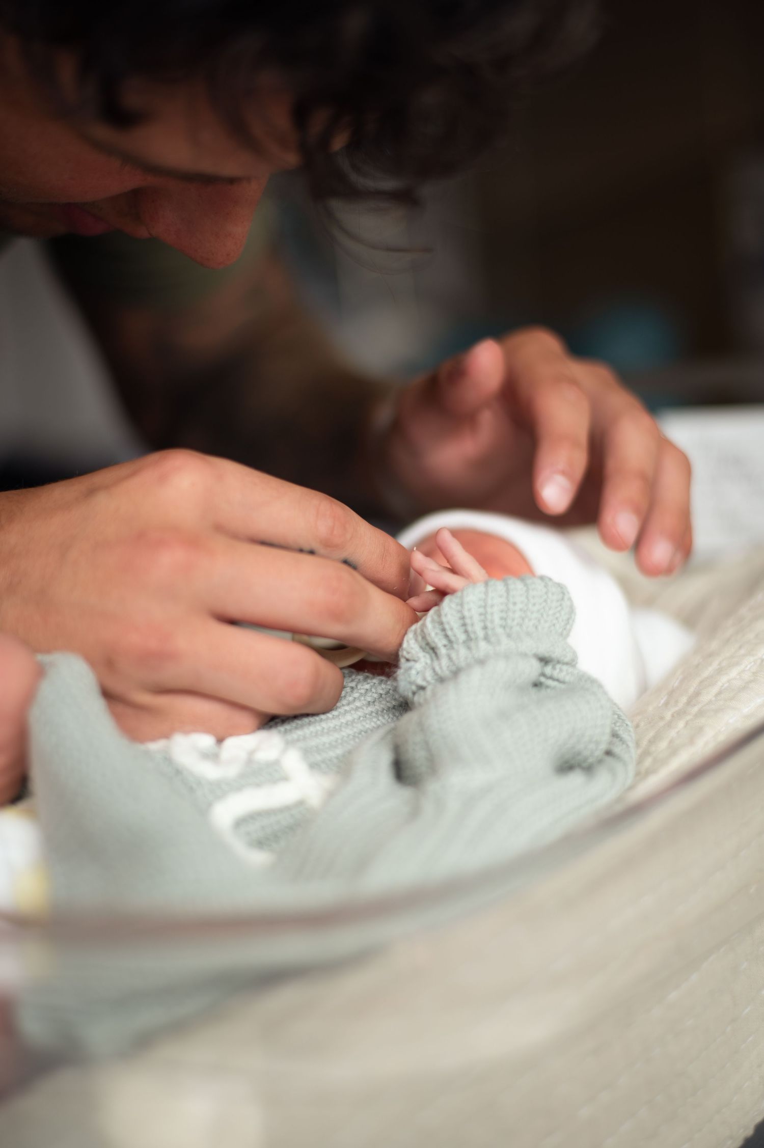 Parent tenderly holding newborn baby's tiny hand, wrapped in soft mint green knit blanket in hospital bassinet.