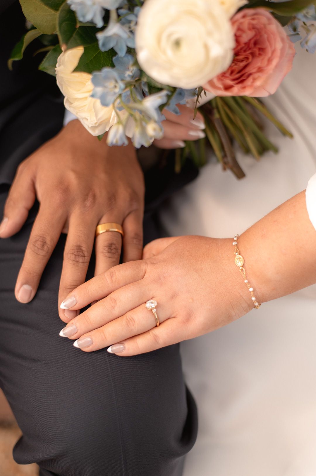 Newlywed couple displaying wedding rings and gold bracelet with a bridal bouquet of pink and blue flowers in the background.