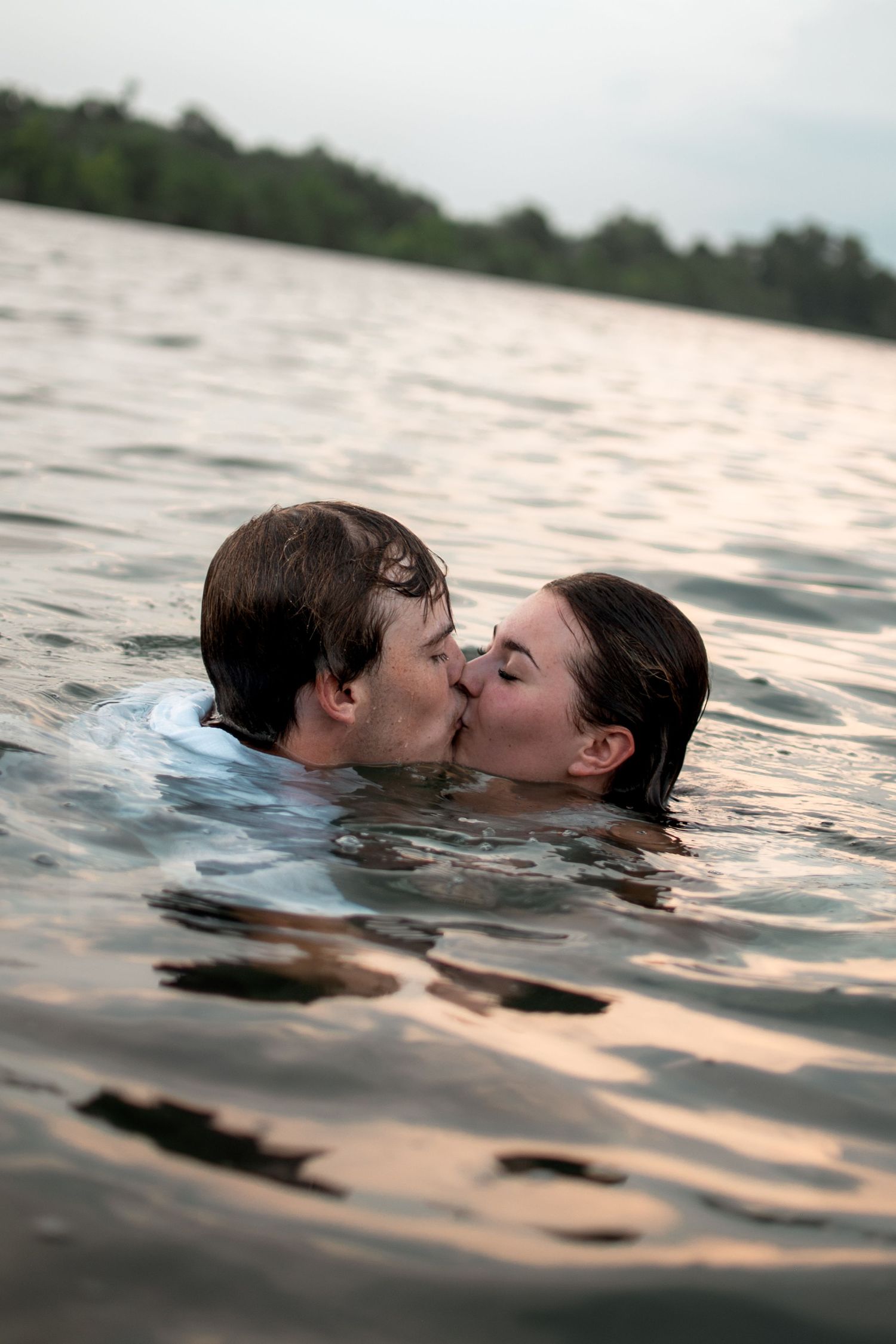 Romantic couple sharing a kiss while swimming in a calm lake at dusk with trees in the background.