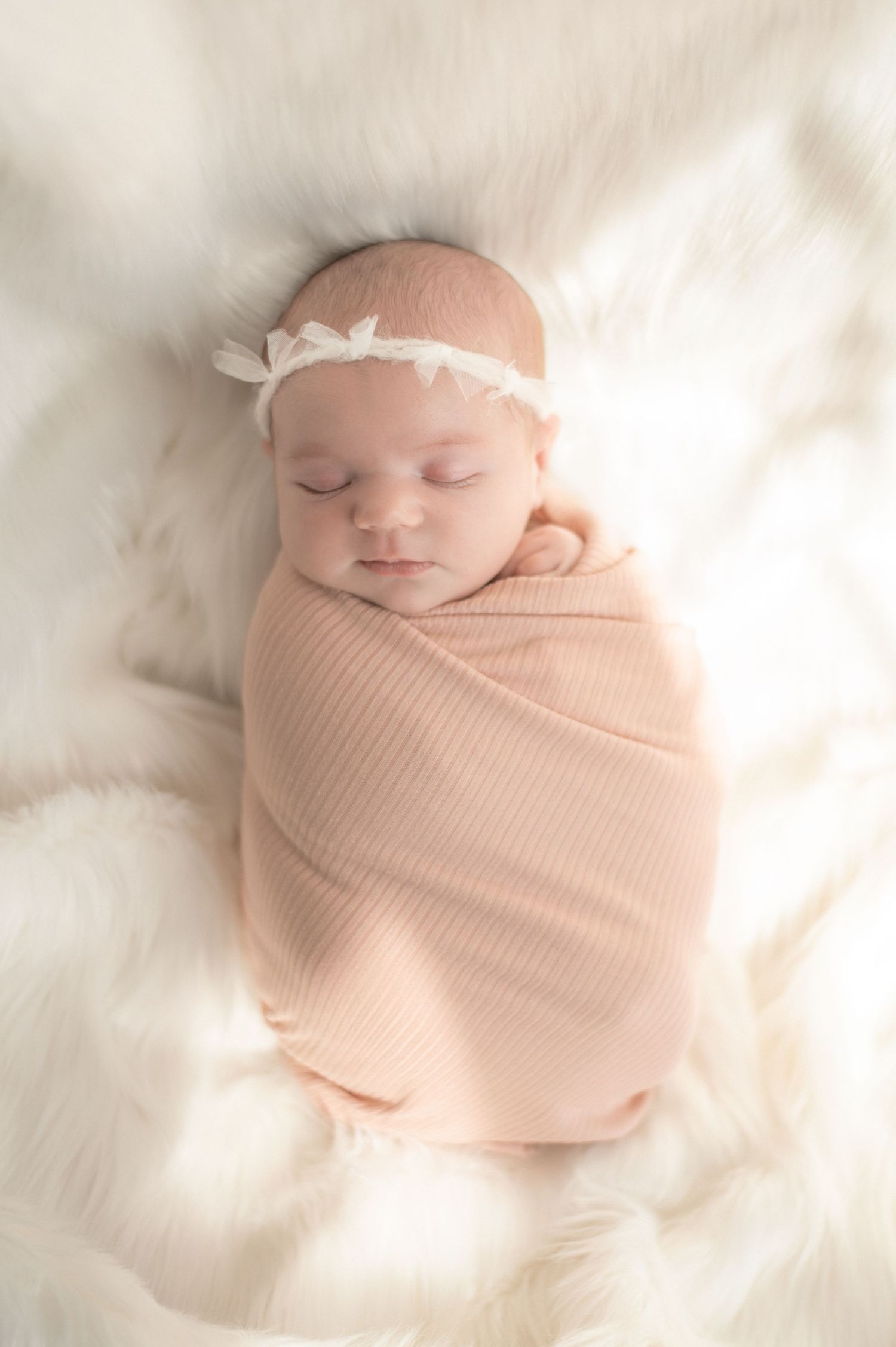 Peaceful newborn baby girl swaddled in a soft peach wrap with a white floral headband, sleeping on a fluffy white blanket.