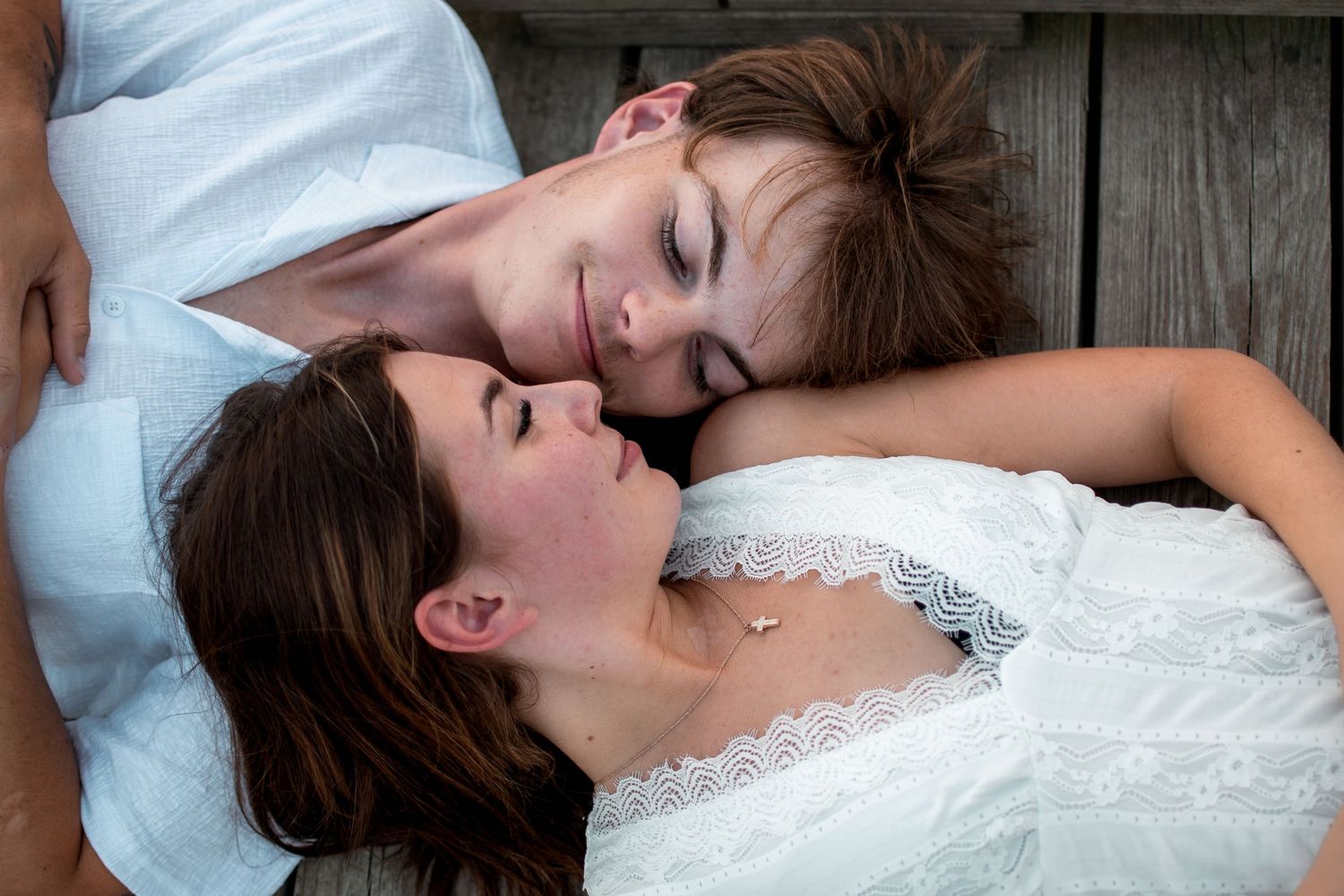 Two young women in white dresses lying on wooden deck, smiling and resting together in a peaceful moment.