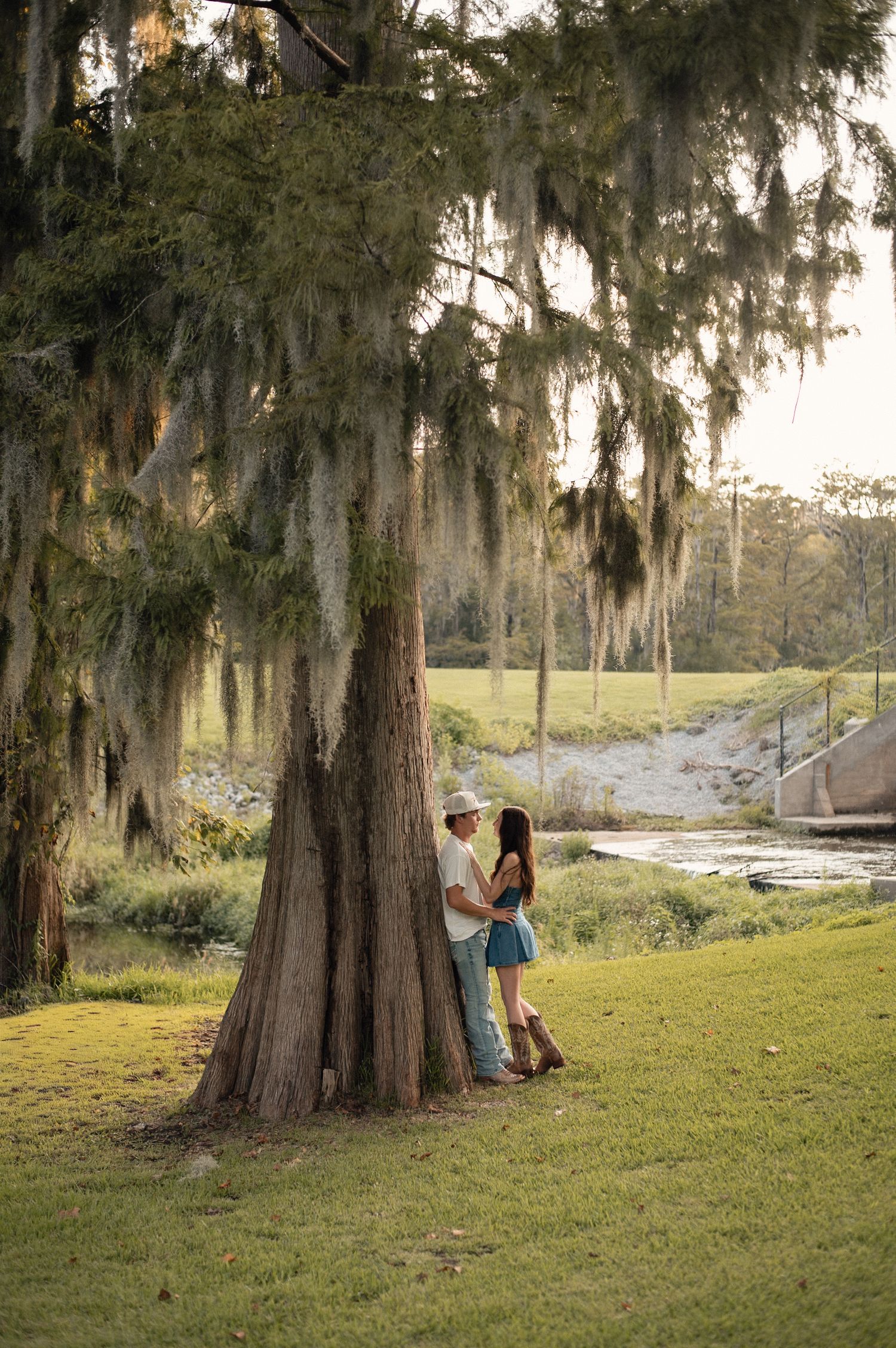 Couple embracing under a large moss-draped tree by a peaceful river in a lush green landscape.