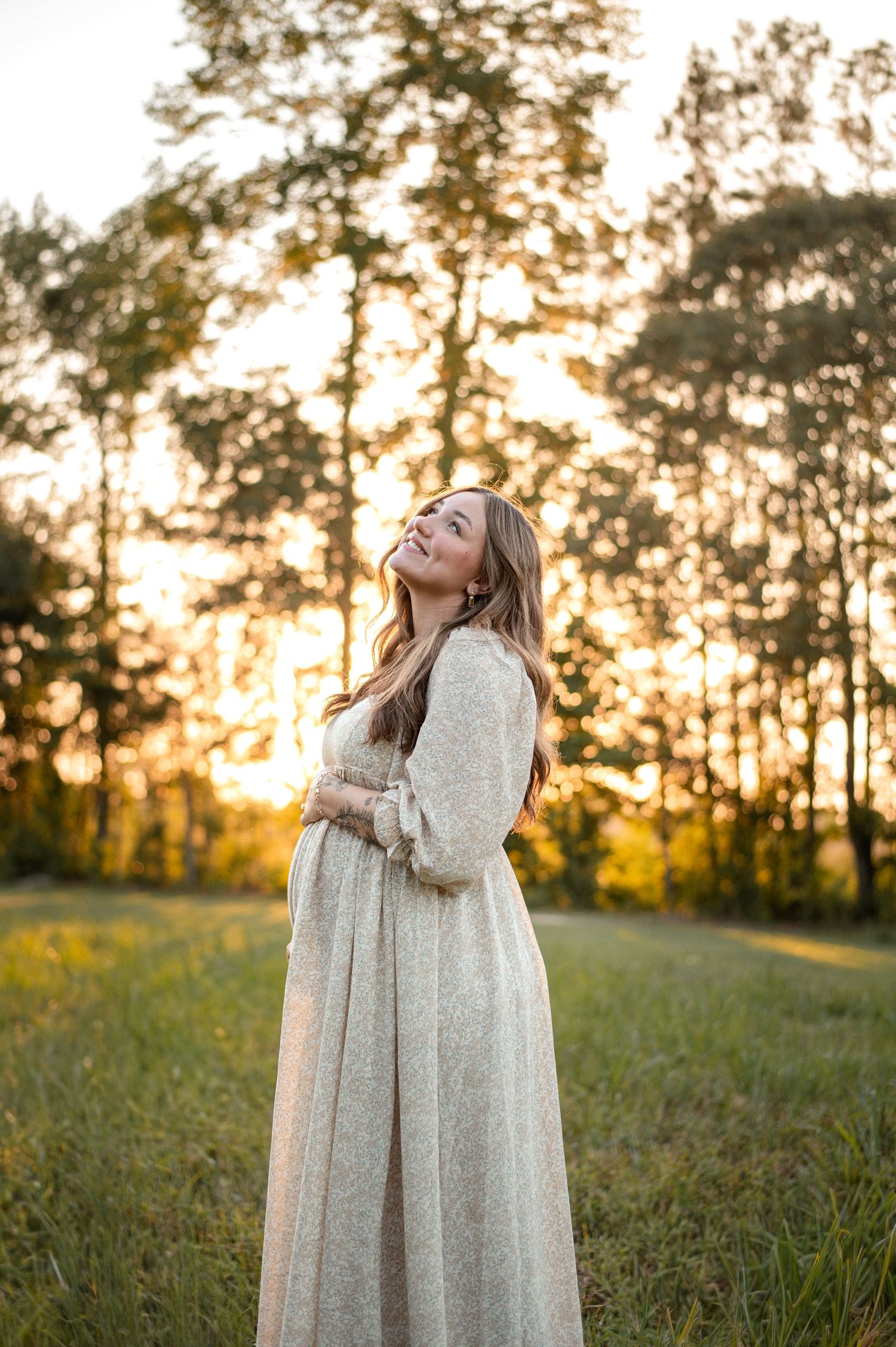 A woman in a flowing white dress stands in a sunlit meadow at golden hour, smiling with eyes closed.