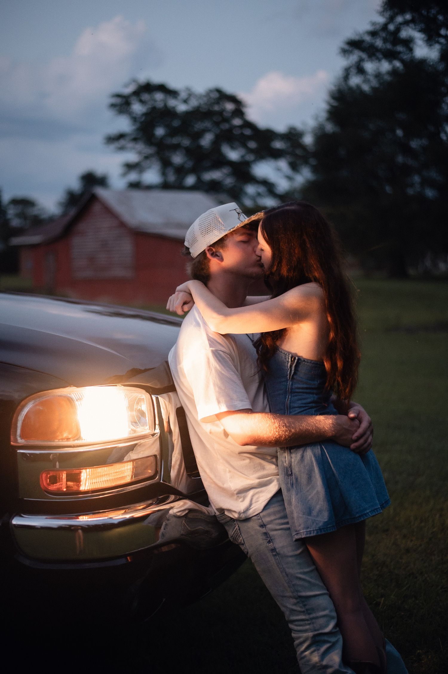 Couple sharing a romantic kiss leaning against a truck at dusk, with a red barn in the background.