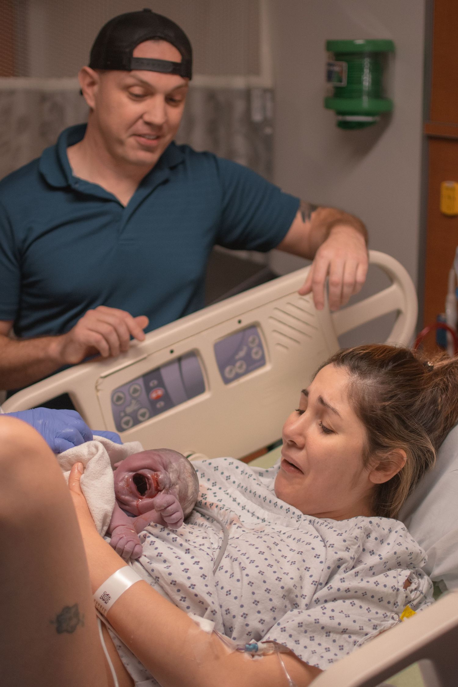 New mother holds newborn baby in hospital bed while father looks on with joy after delivery.