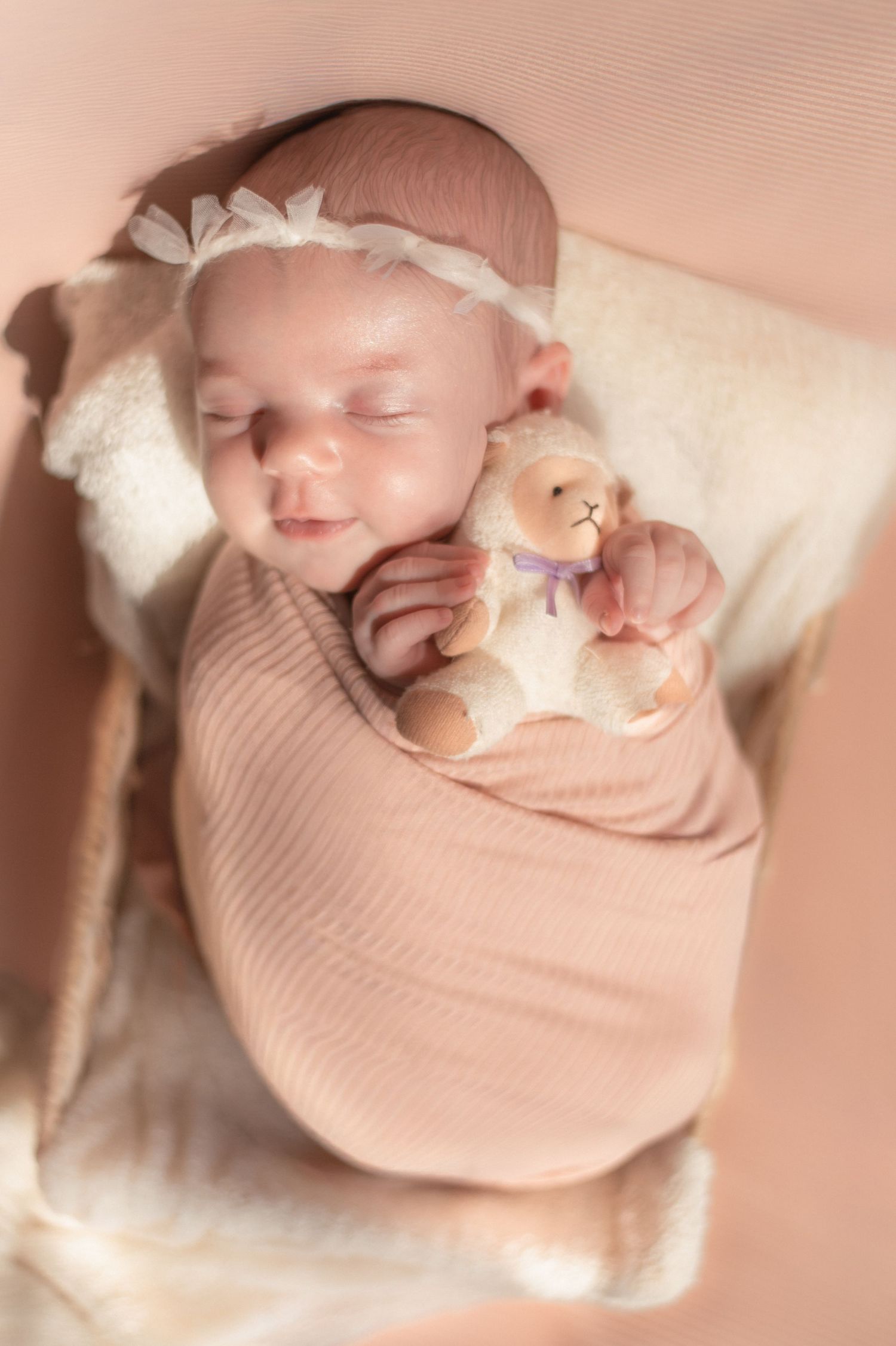 Sleeping newborn baby girl swaddled in pink wrap holding a tiny teddy bear, wearing a white bow headband.