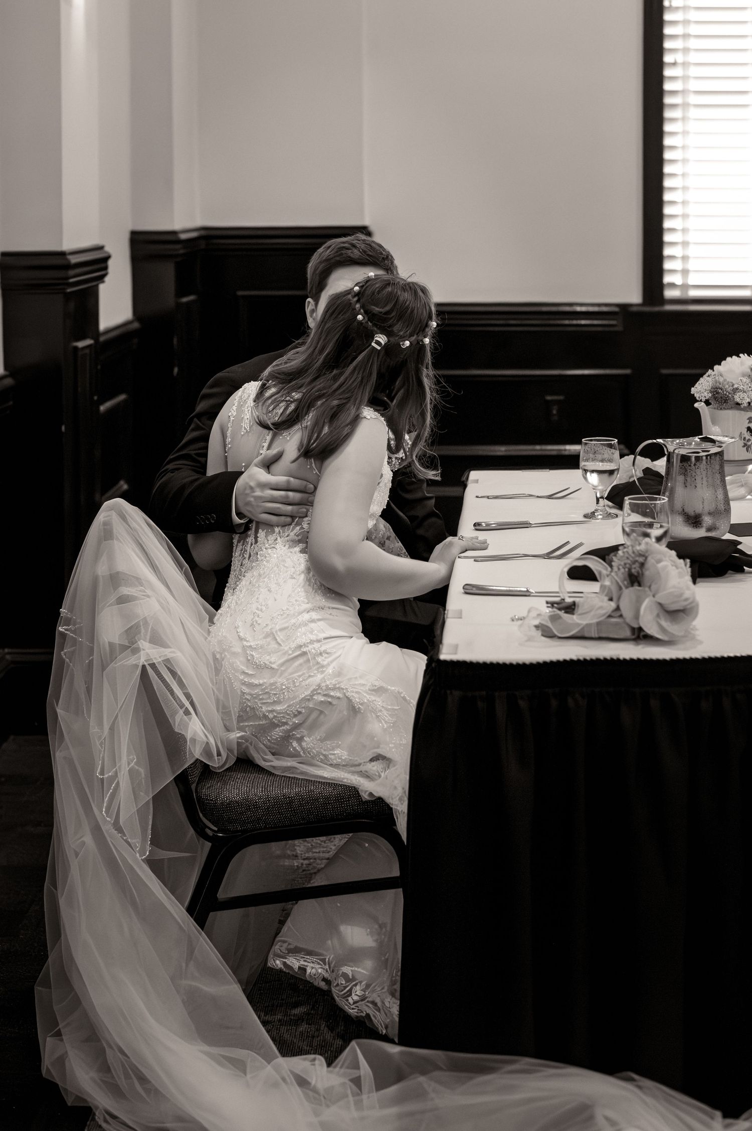 Black and white photo of a bride and groom sharing a kiss at their wedding reception table, with bridal veil flowing.