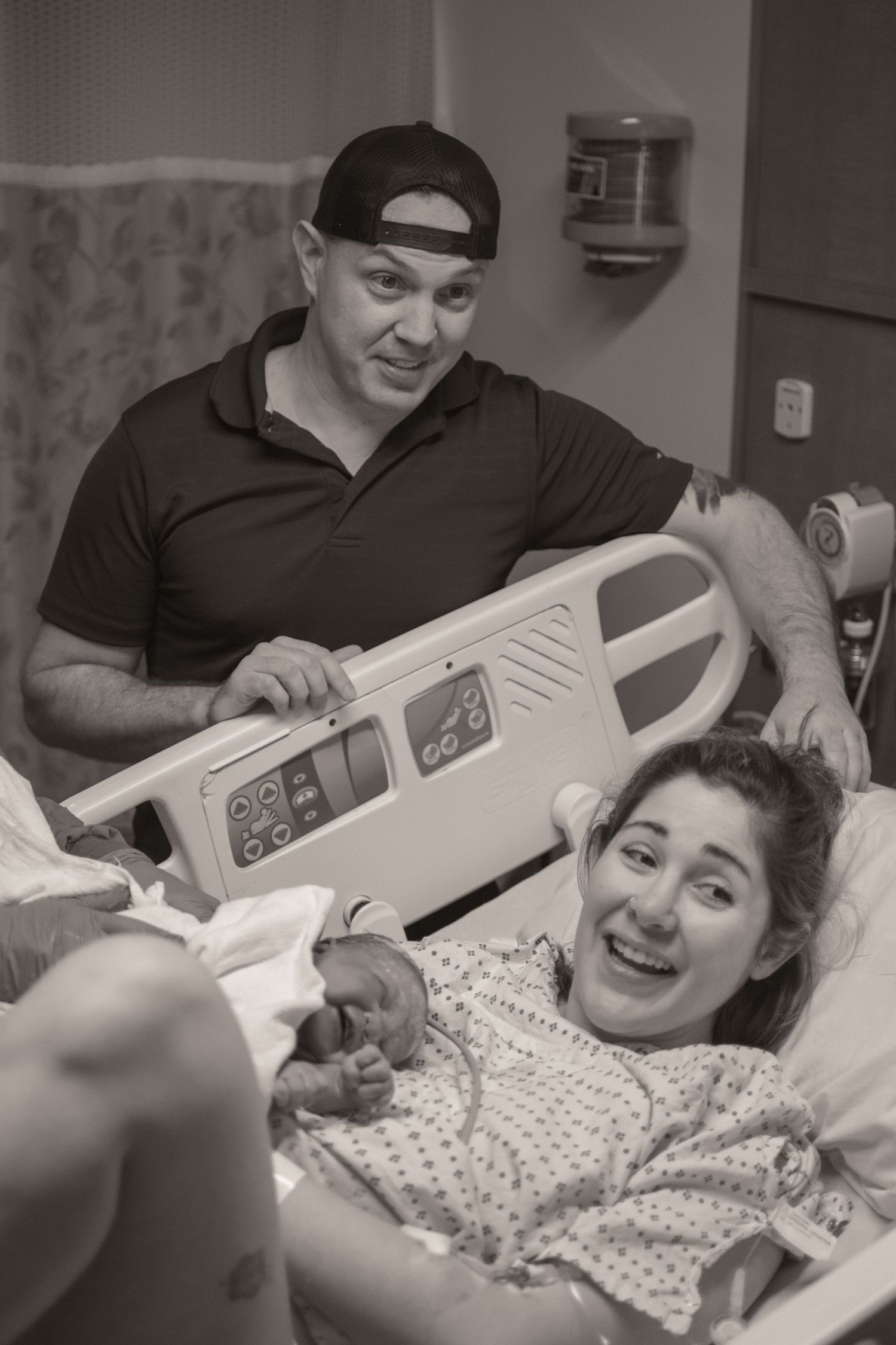 Black and white photo of smiling new parents in hospital room, mother holding newborn baby in bed after delivery.
