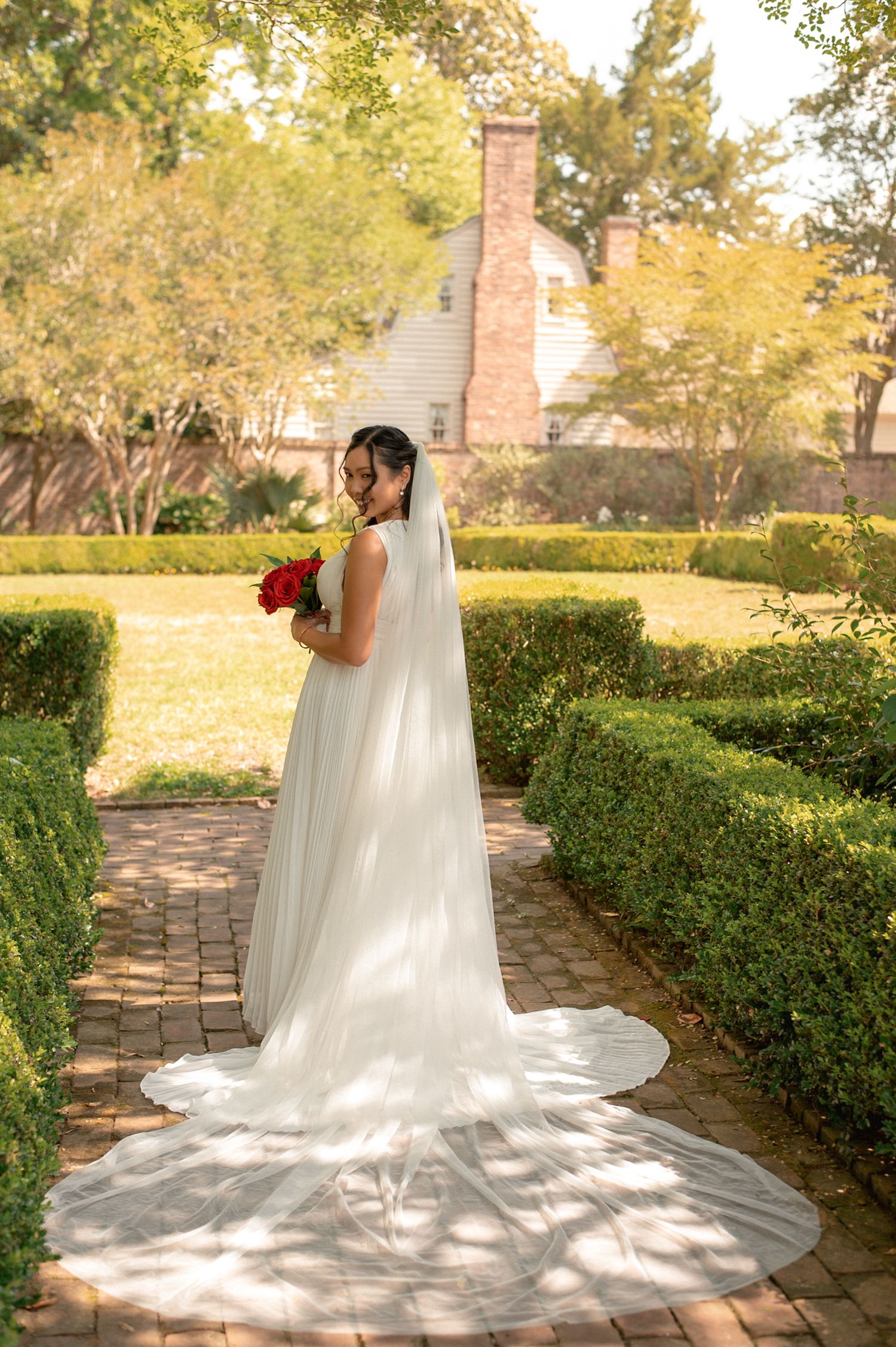 Bride in white gown and veil holds red bouquet on brick path in lush garden near brick house.