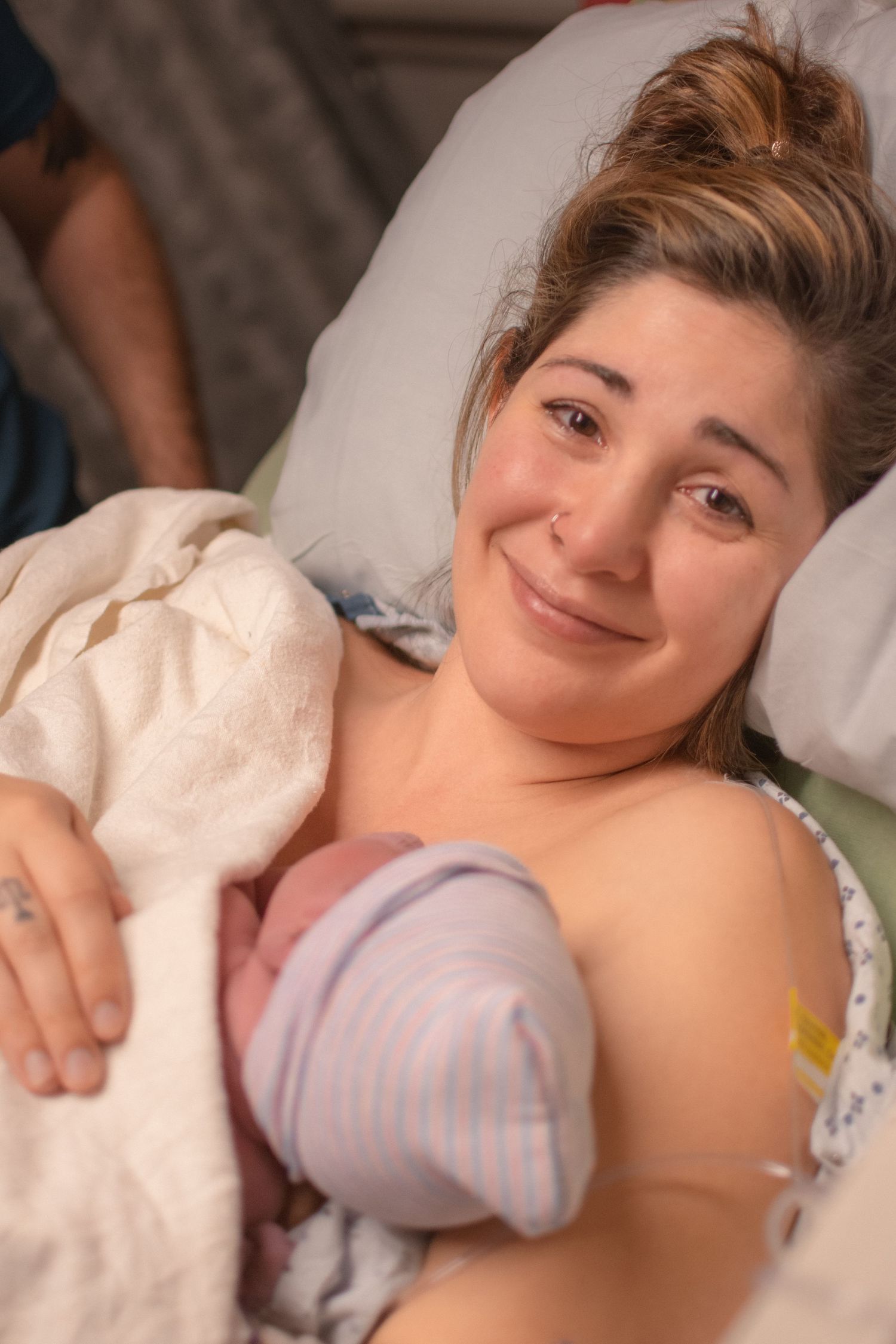 New mother smiling warmly in hospital bed while holding her newborn baby wrapped in a striped blanket after delivery.