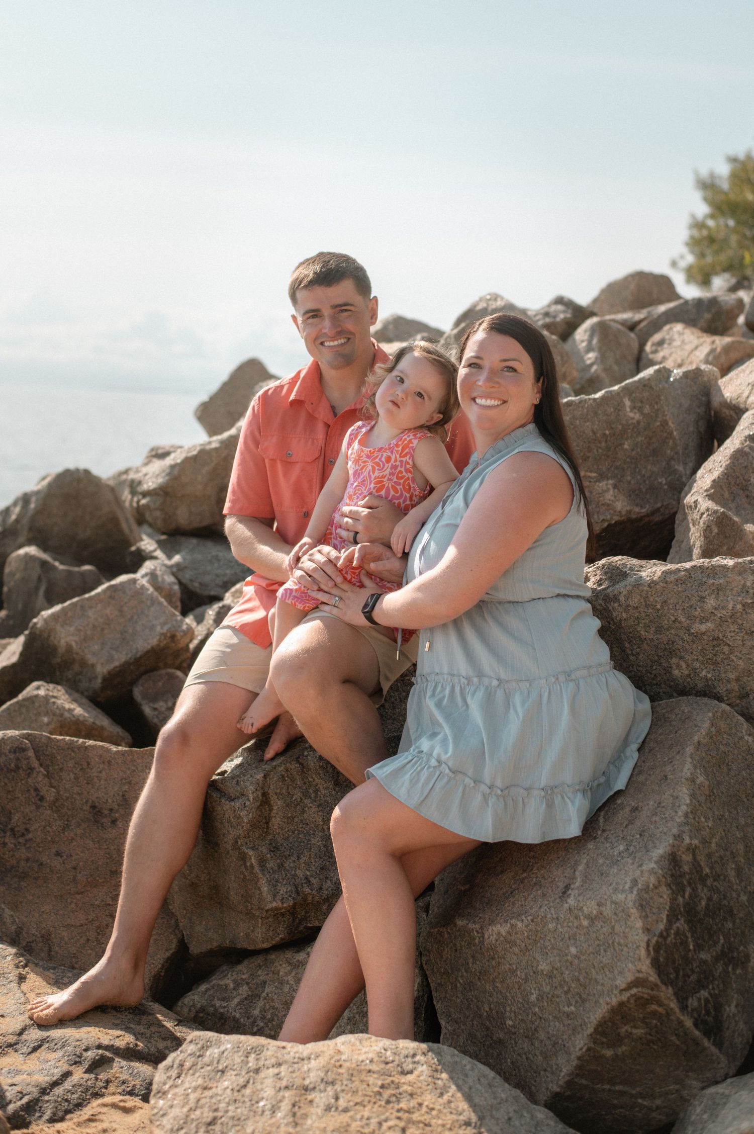 Happy family of three sitting on rocky shore, parents smiling with young child, ocean backdrop on sunny day.