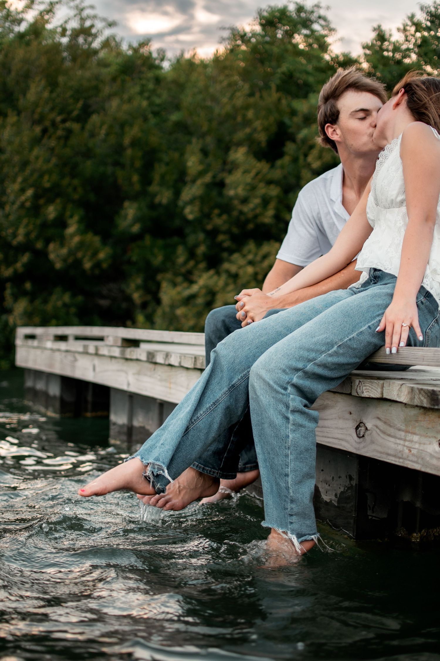Couple kissing on wooden dock with feet in water, wearing jeans, surrounded by lush green trees at dusk.