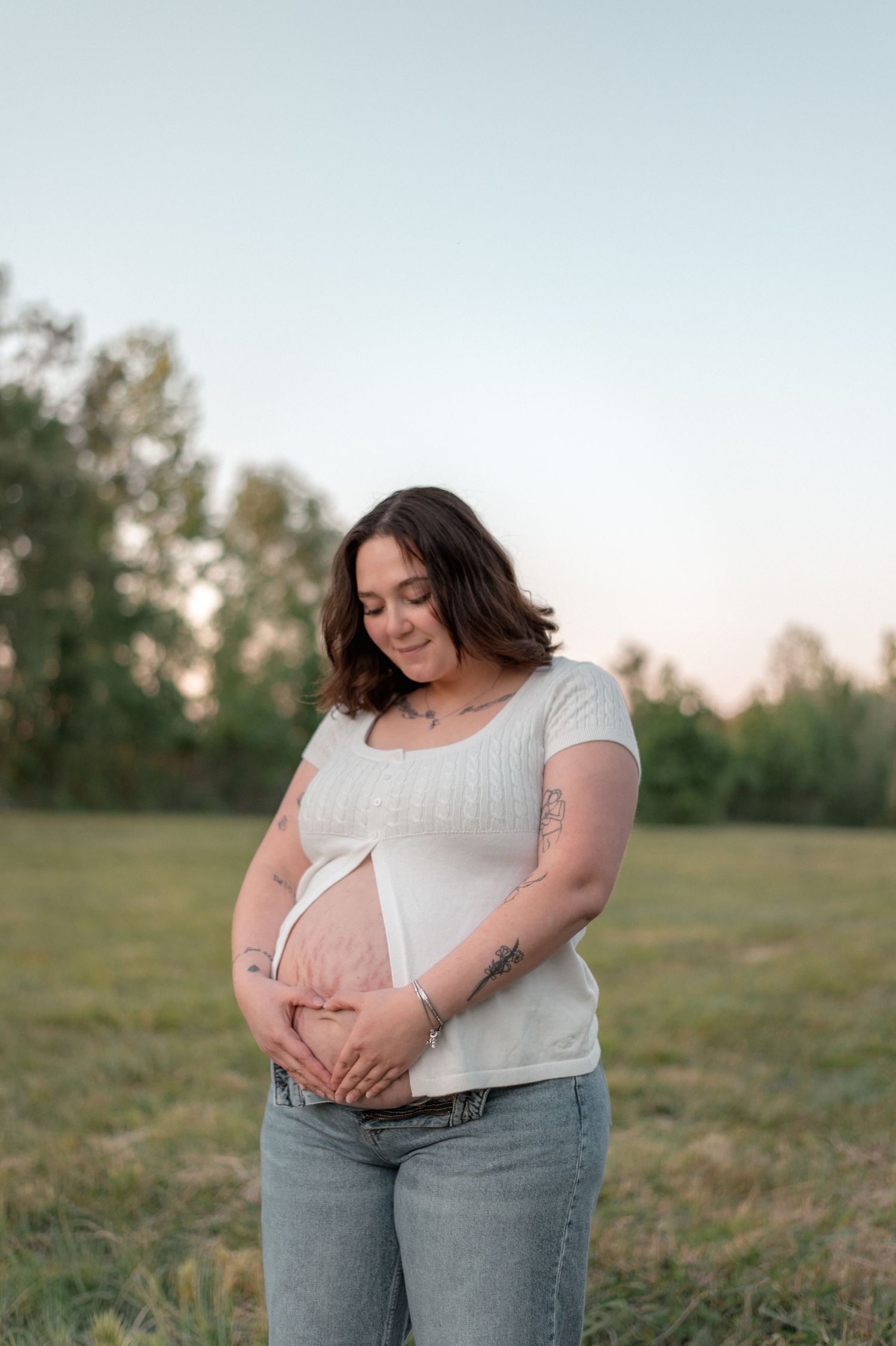 Pregnant woman in white top and jeans cradling her baby bump in a green field at golden hour.