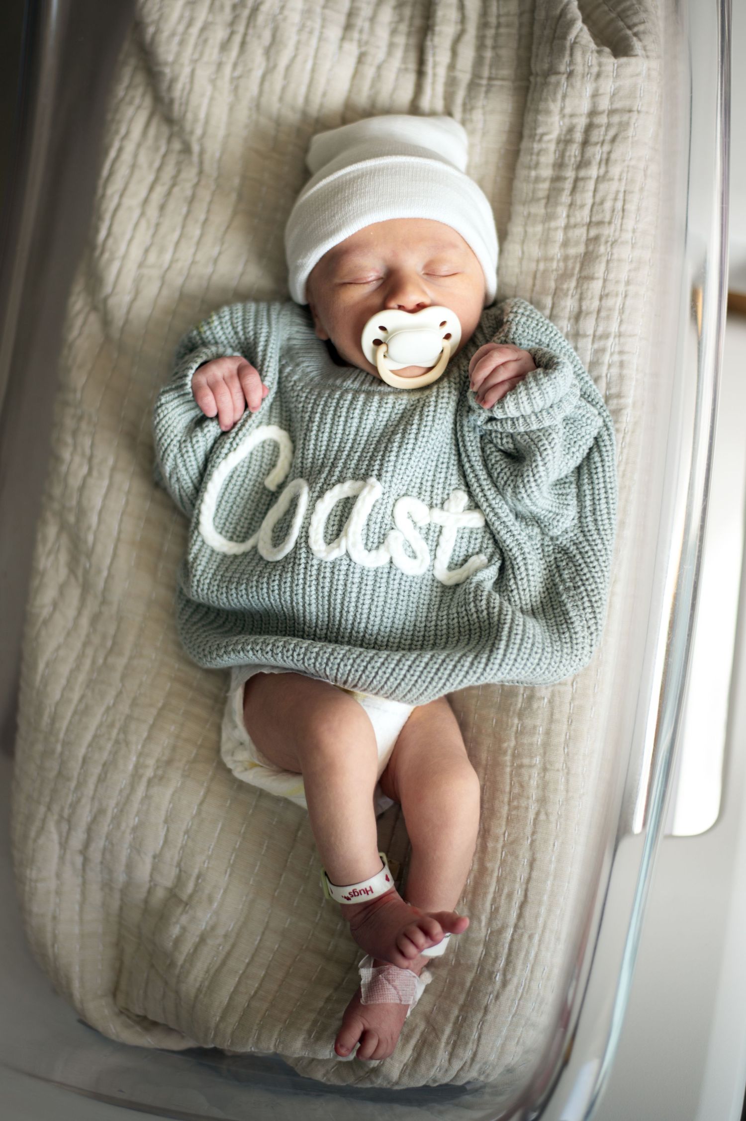 Newborn baby named Coast wearing a sage green knit sweater and white hat, sleeping peacefully with a pacifier.