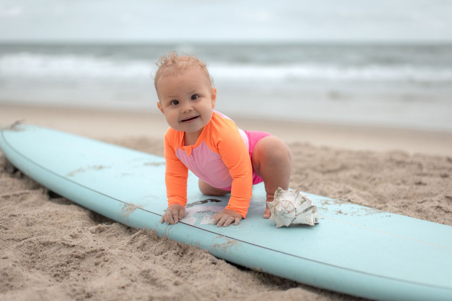 Baby in pink swimsuit smiling on a surfboard at the beach.