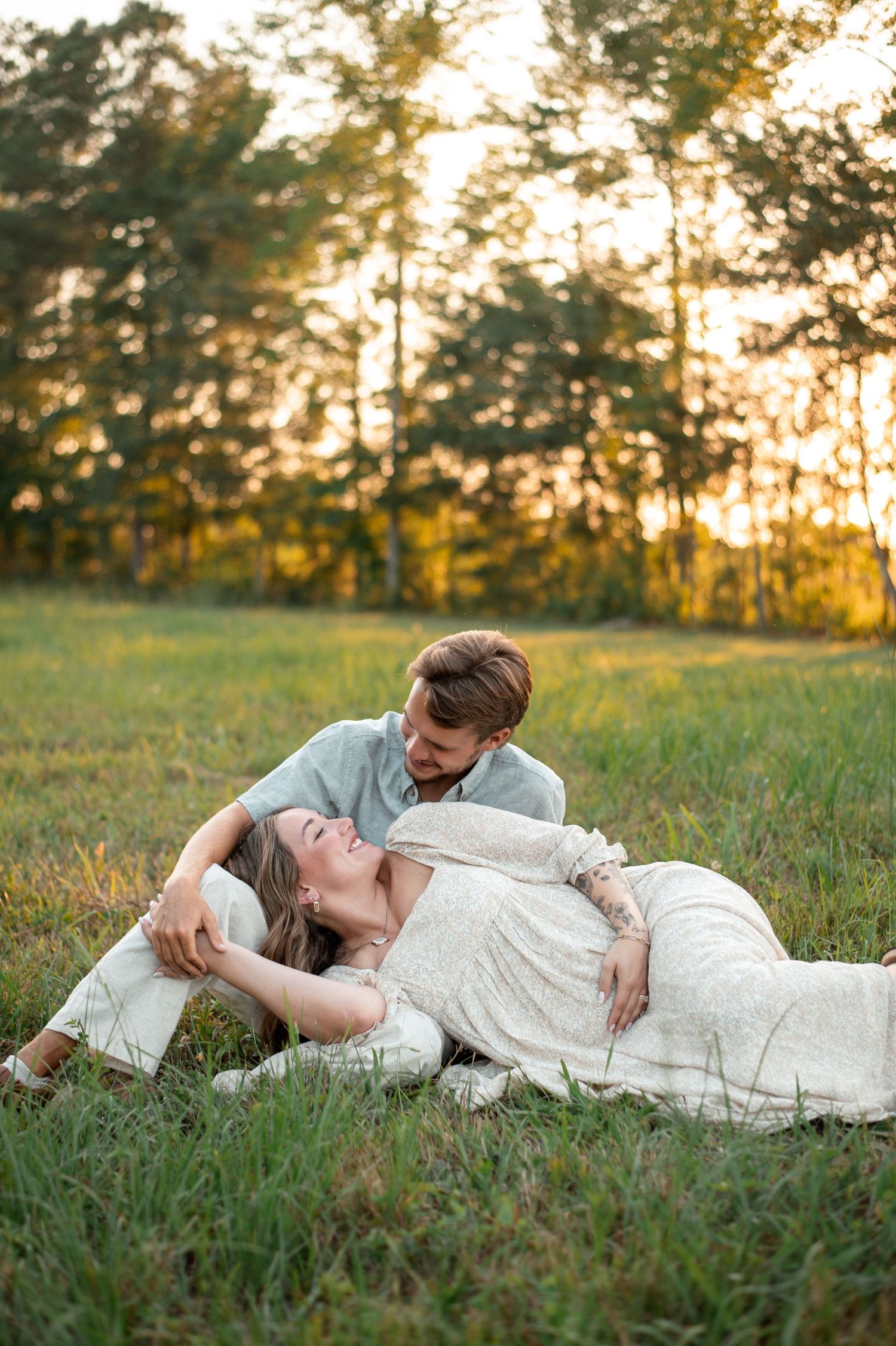 A couple shares a tender moment in a sunlit meadow, the woman lying down smiling as the man gazes at her lovingly.