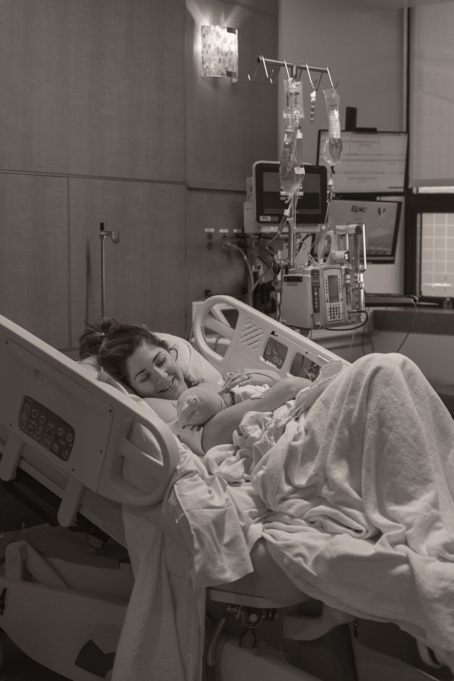 New mother resting in hospital bed holding newborn baby after childbirth, black and white photo.