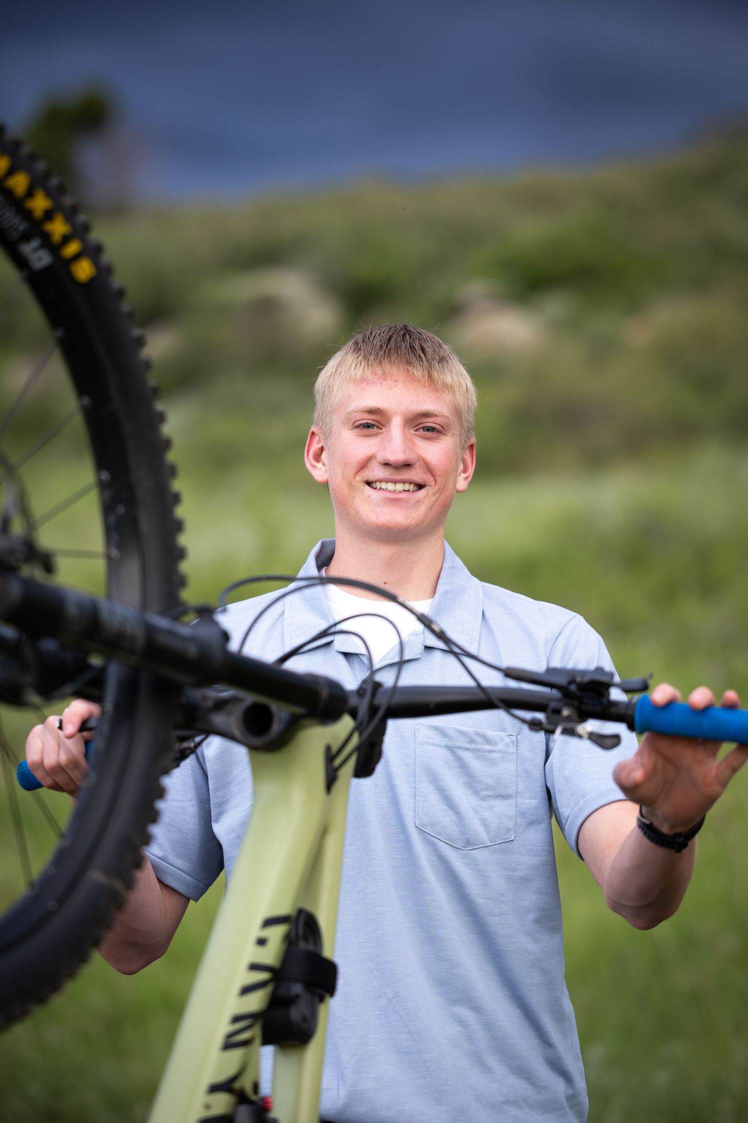 Smiling blonde teen holds mountain bike upright on green hillside in outdoor portrait photo.