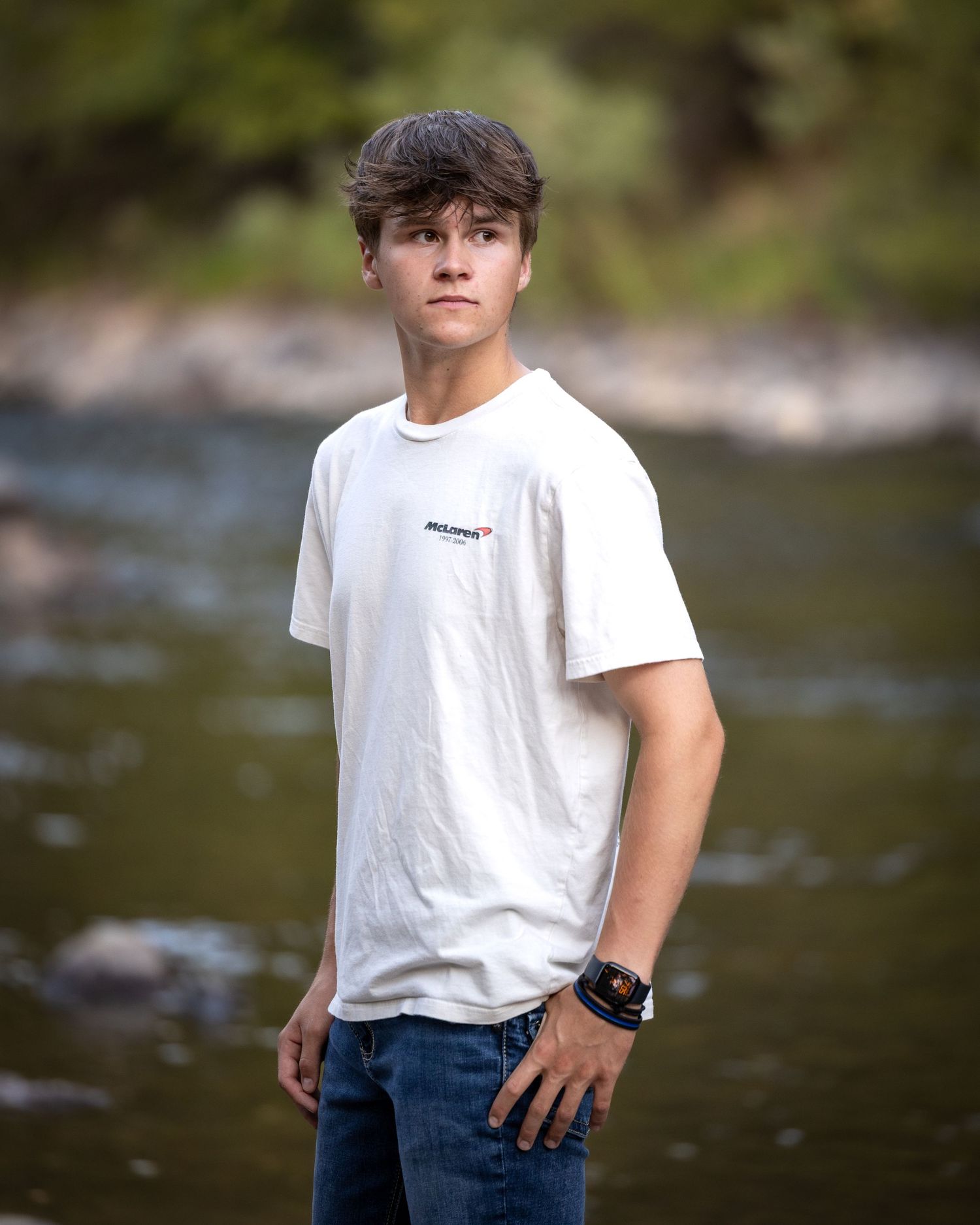 Teen boy in white McLaren t-shirt stands confidently before a rocky river backdrop outdoors.