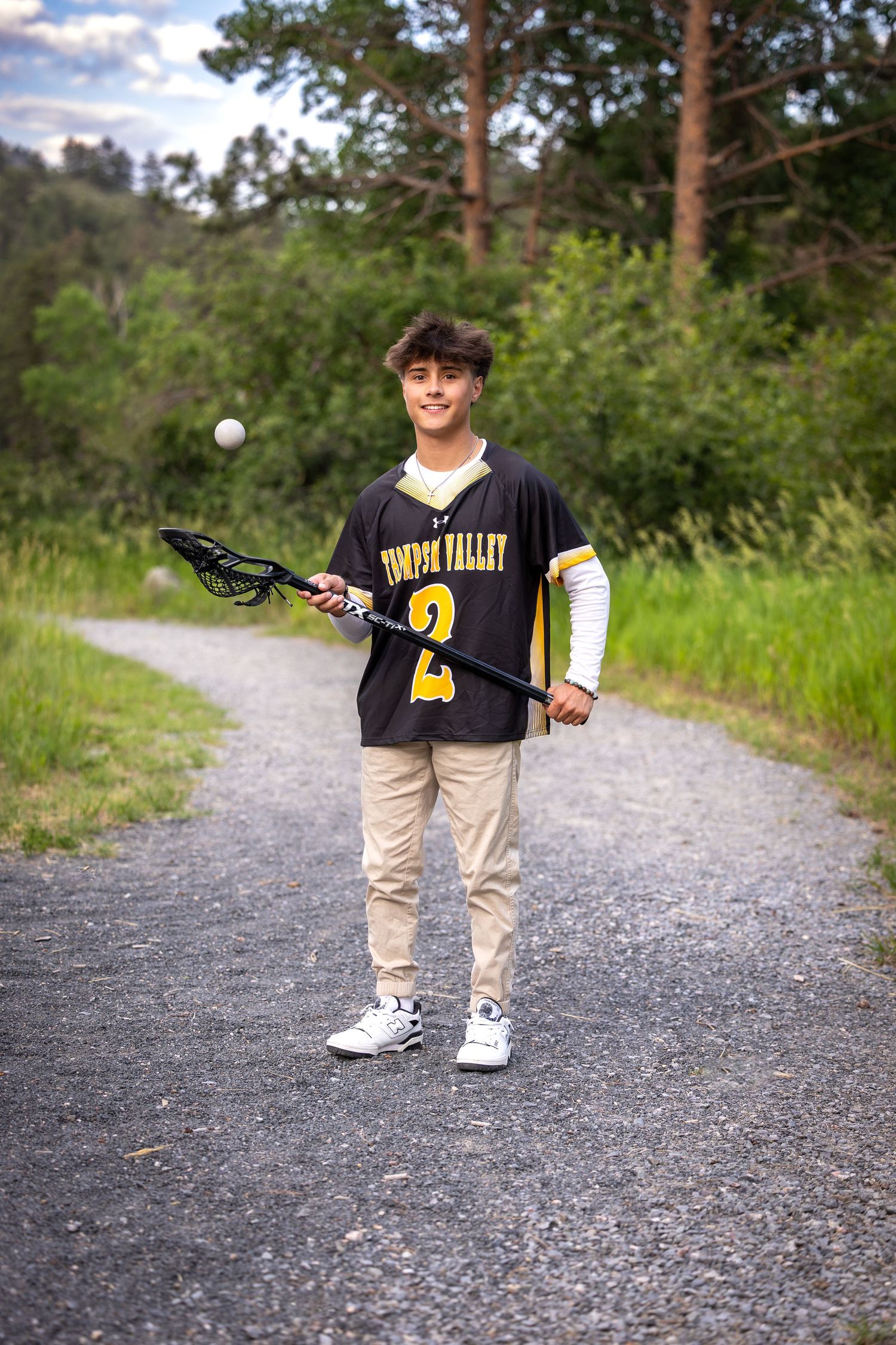 Teen lacrosse player in Thompson Valley jersey holds stick on gravel path surrounded by greenery.