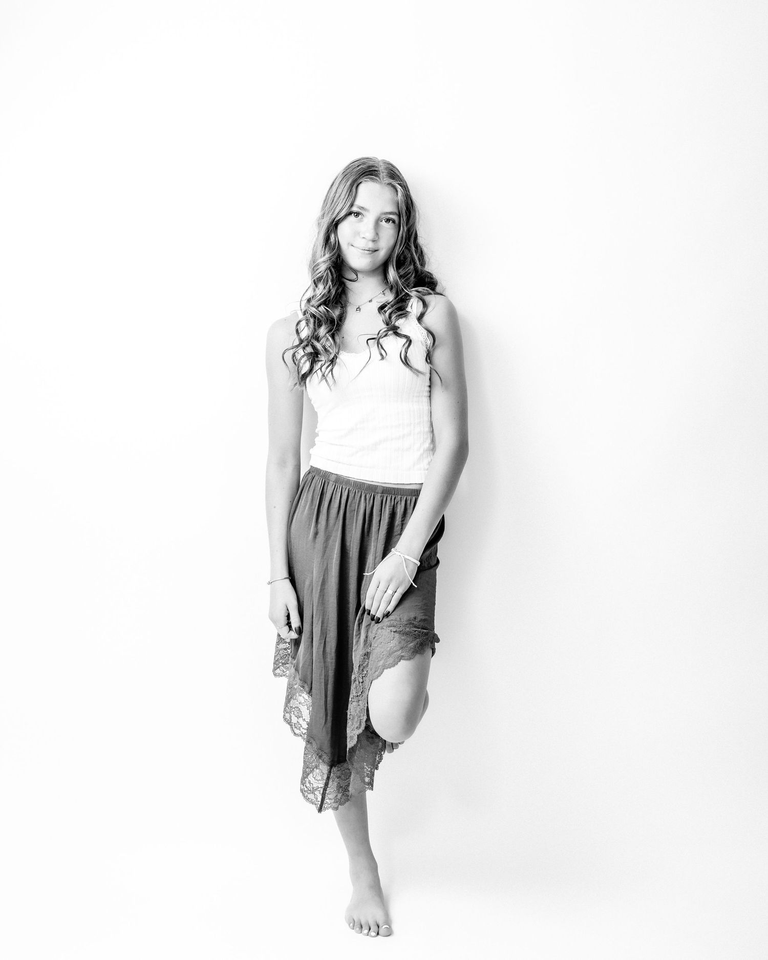 Young woman with long curly hair stands barefoot in a white tank and flowy skirt in a black and white portrait.