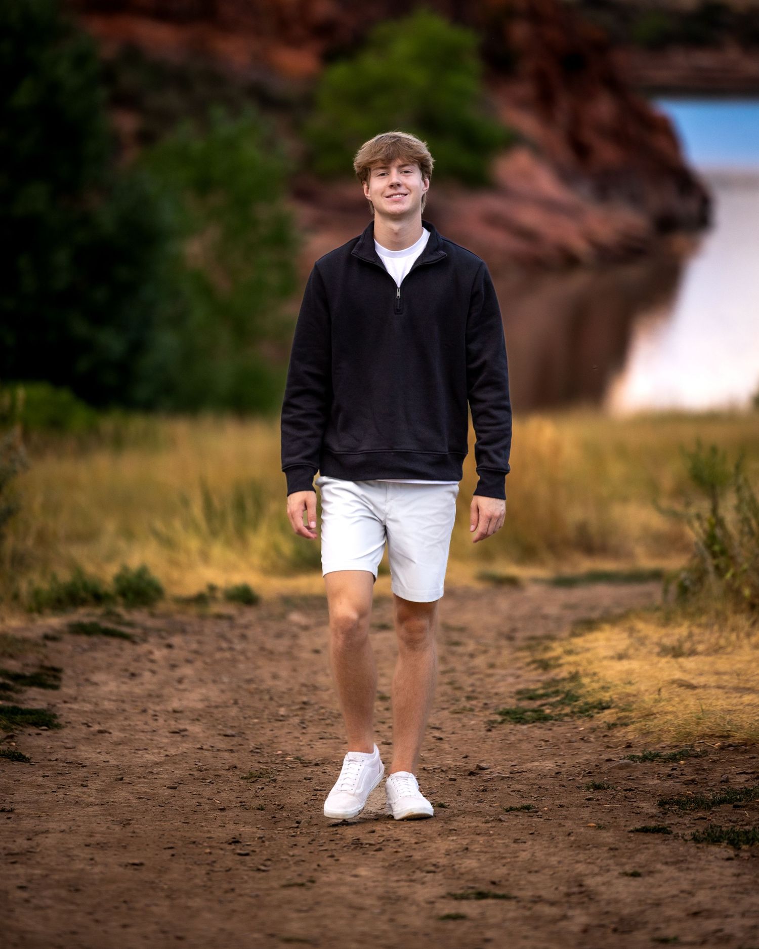 Teen boy in black hoodie and white shorts walking on dirt path near red rock formations.