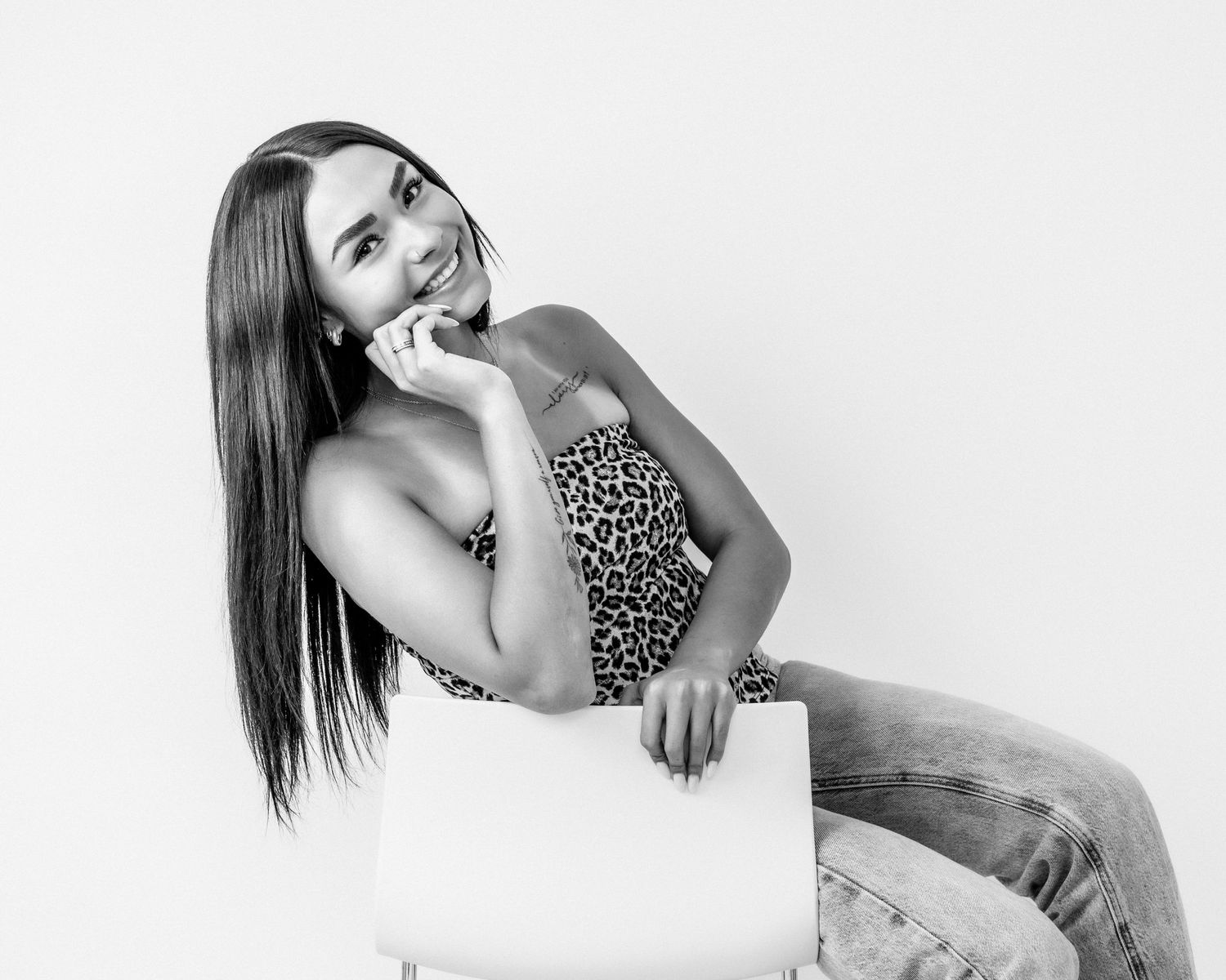 Young woman with straight dark hair sits on a white chair smiling in a leopard print top in a studio portrait.