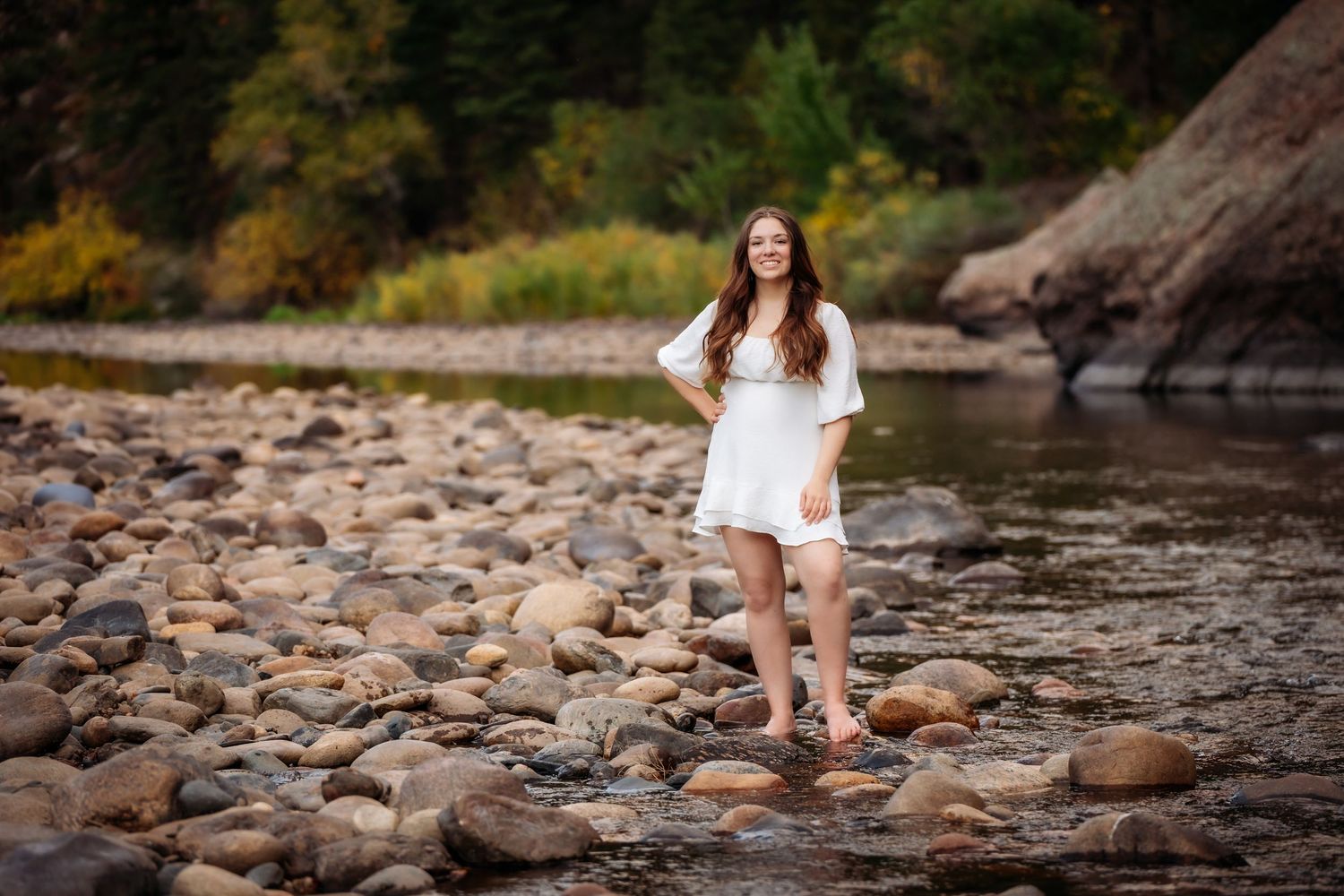 Young woman in white dress standing barefoot on rocky riverbank surrounded by fall colors.