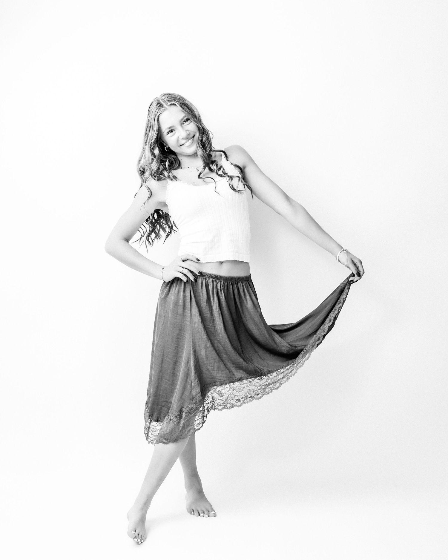 Black and white photo of a smiling girl in a white top and flowing midi skirt posing barefoot.