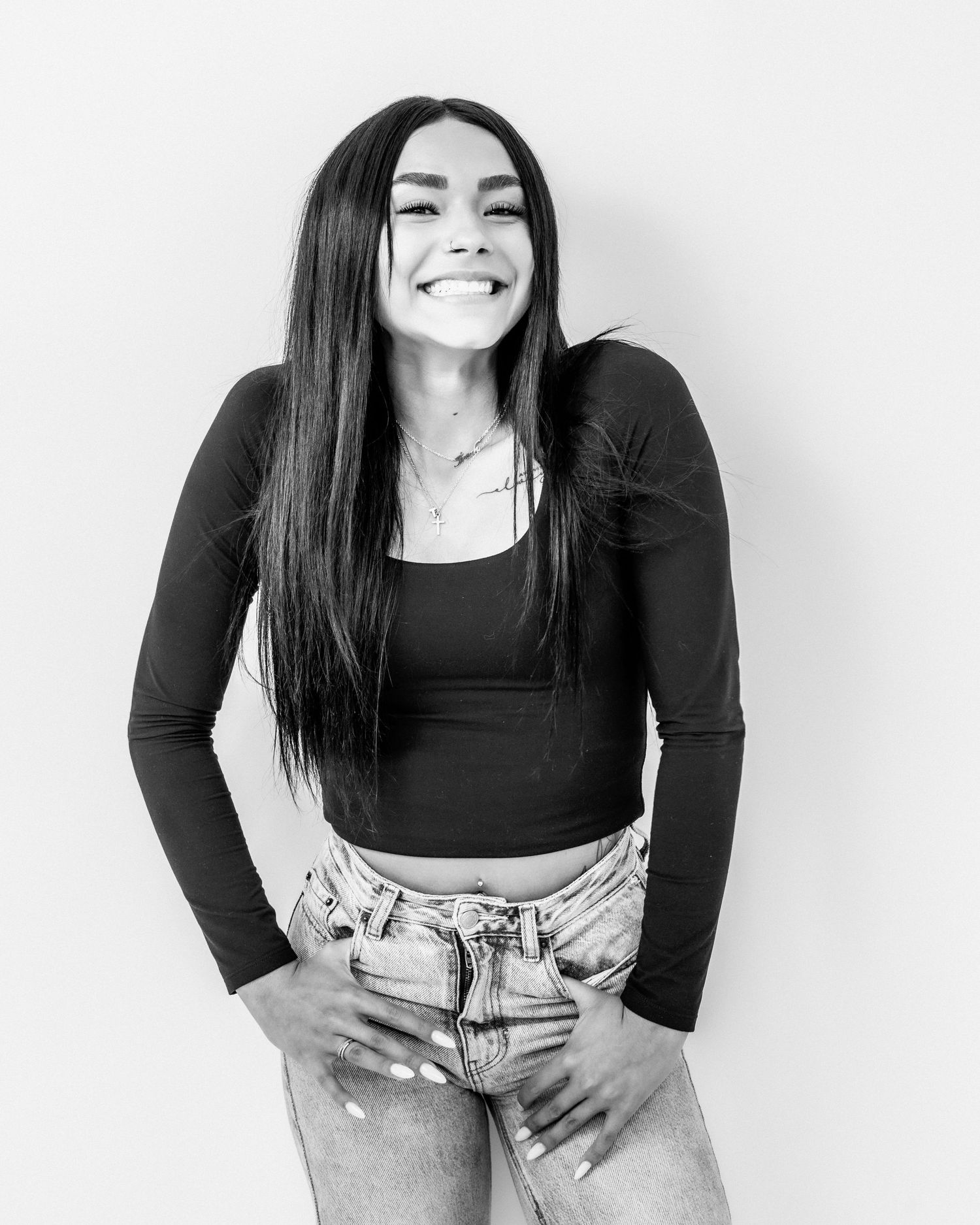 Woman with long dark hair laughs with hands on hips wearing a black top and jeans in a studio portrait.